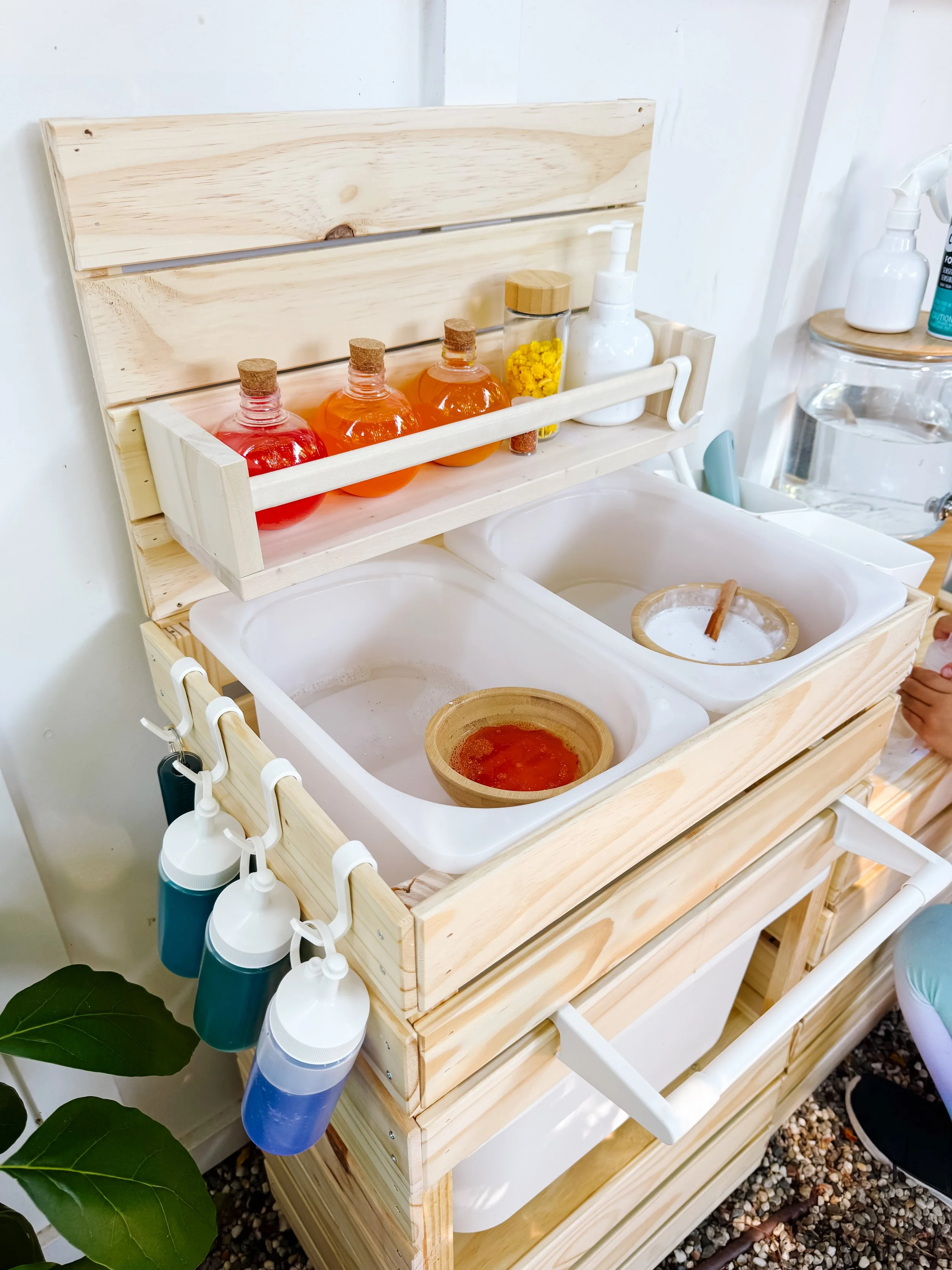 Mud kitchen setup with colored water, bowls, brushes, bottles, and pretend recipe supplies.