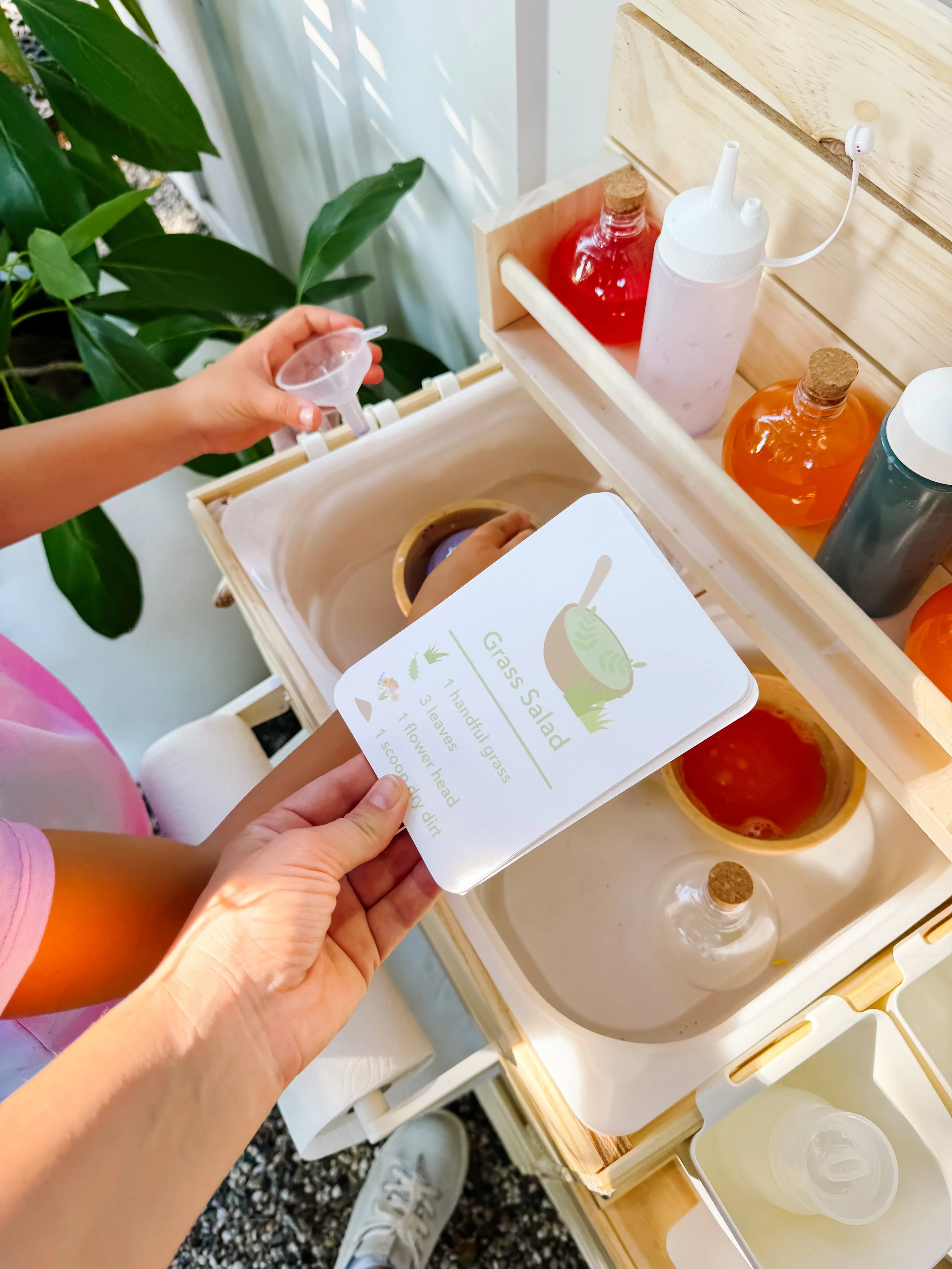 Child using a grass salad recipe card in a mud kitchen with colored water and bowls.