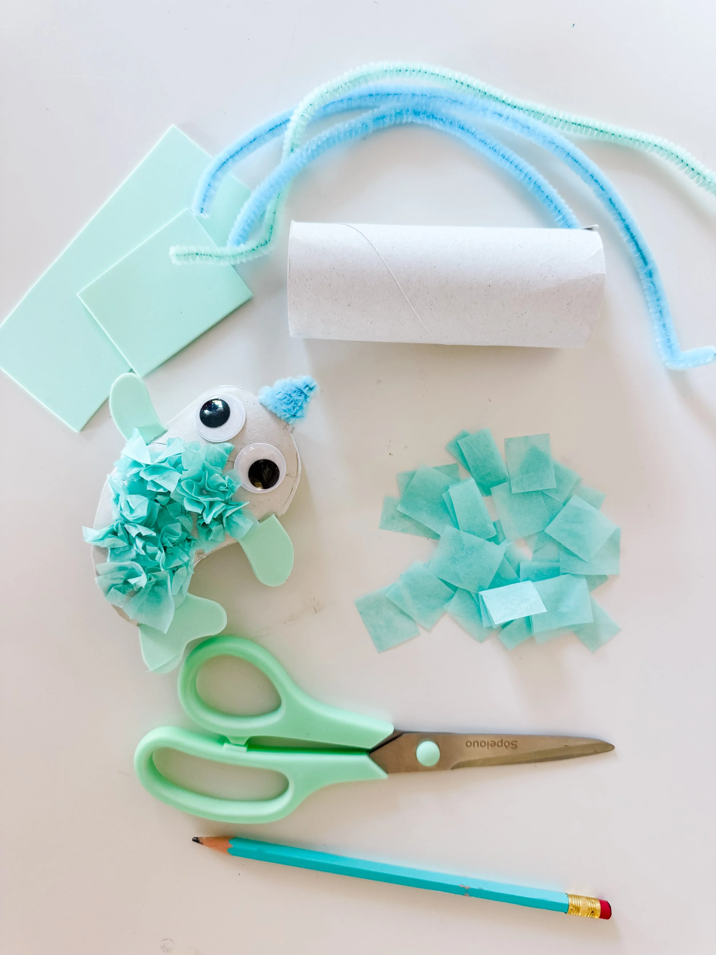Supplies for making a toilet roll narwhal craft including tissue paper, pipe cleaners, scissors, pencil, foam, and googly eyes.