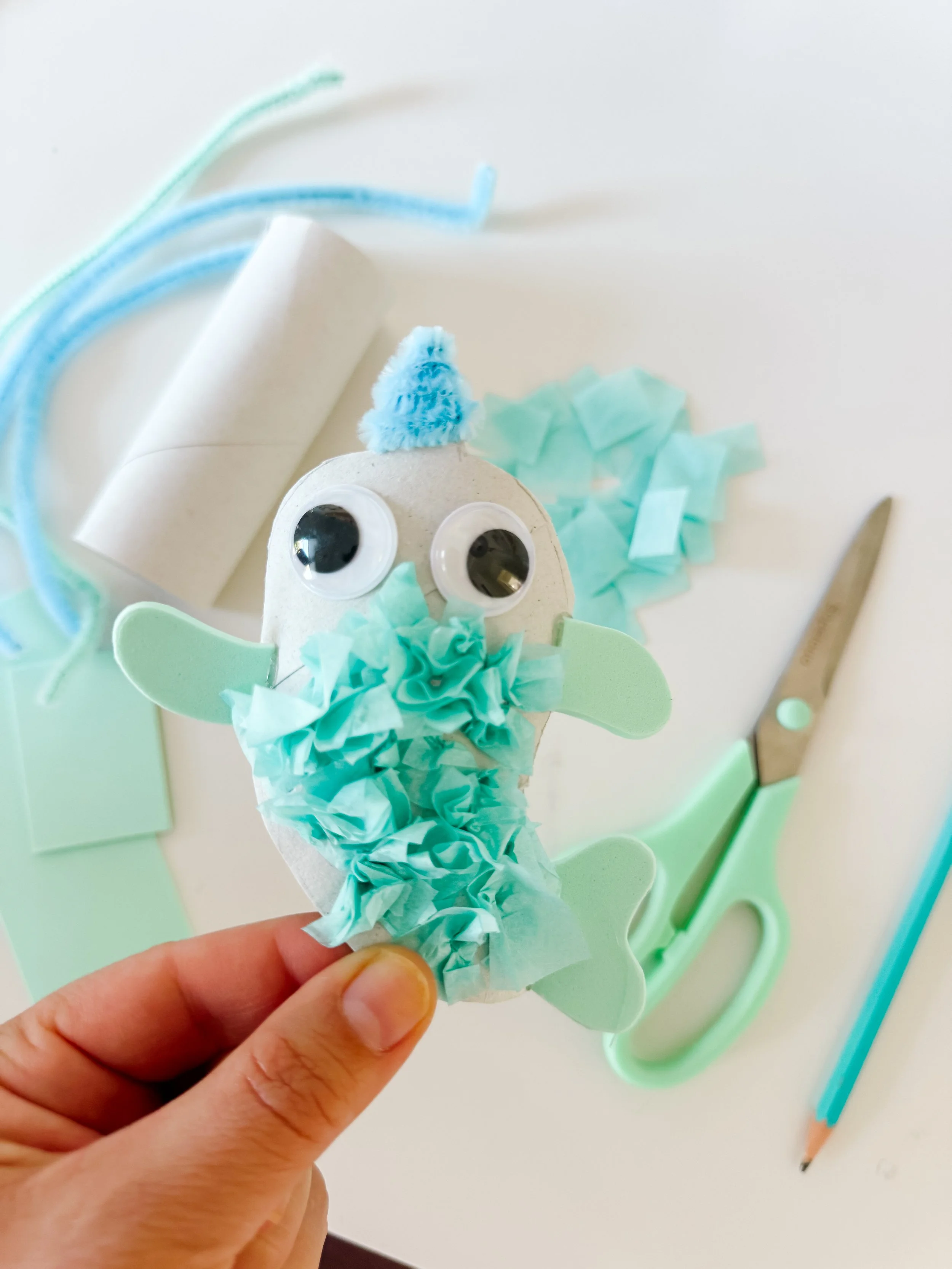 Finished toilet roll narwhal craft with tissue paper body, blue pipe cleaner horn, foam fins, and googly eyes.