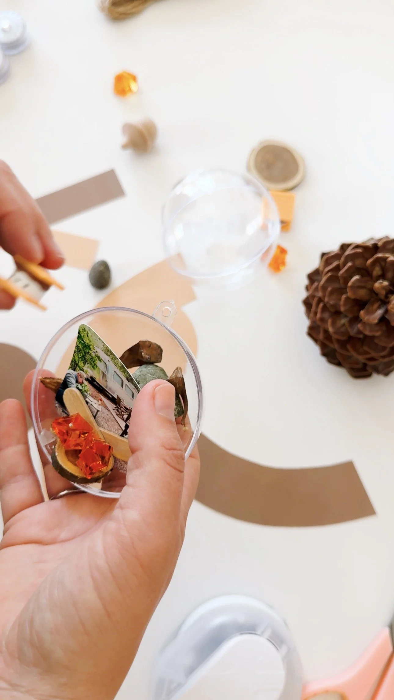 Placing a miniature camping scene with photo, rocks, bark, and glowing orange gems inside a clear plastic ornament.