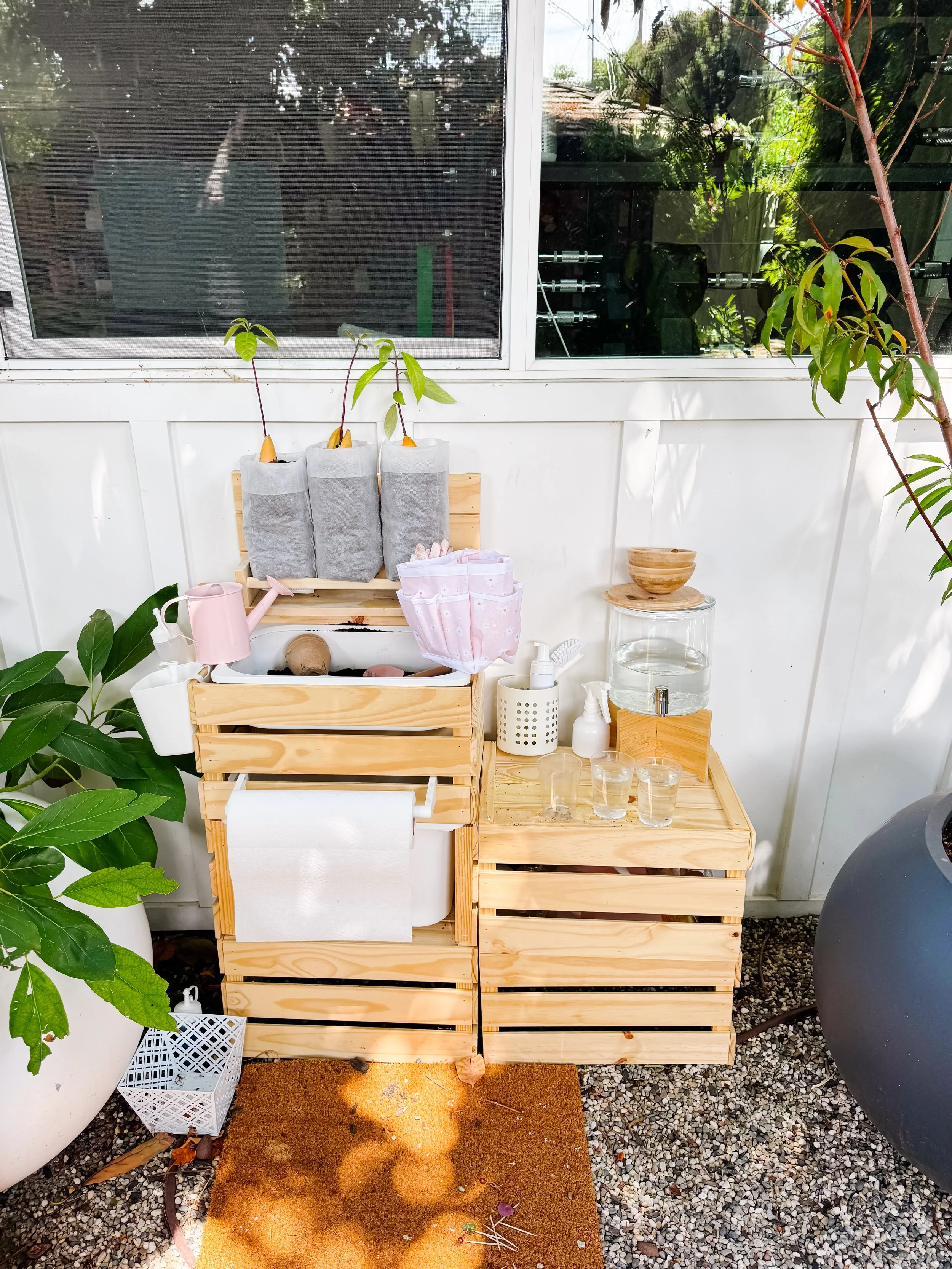 Outdoor mud kitchen garden station with avocado seedlings, watering tools, soil and clear observation glasses.