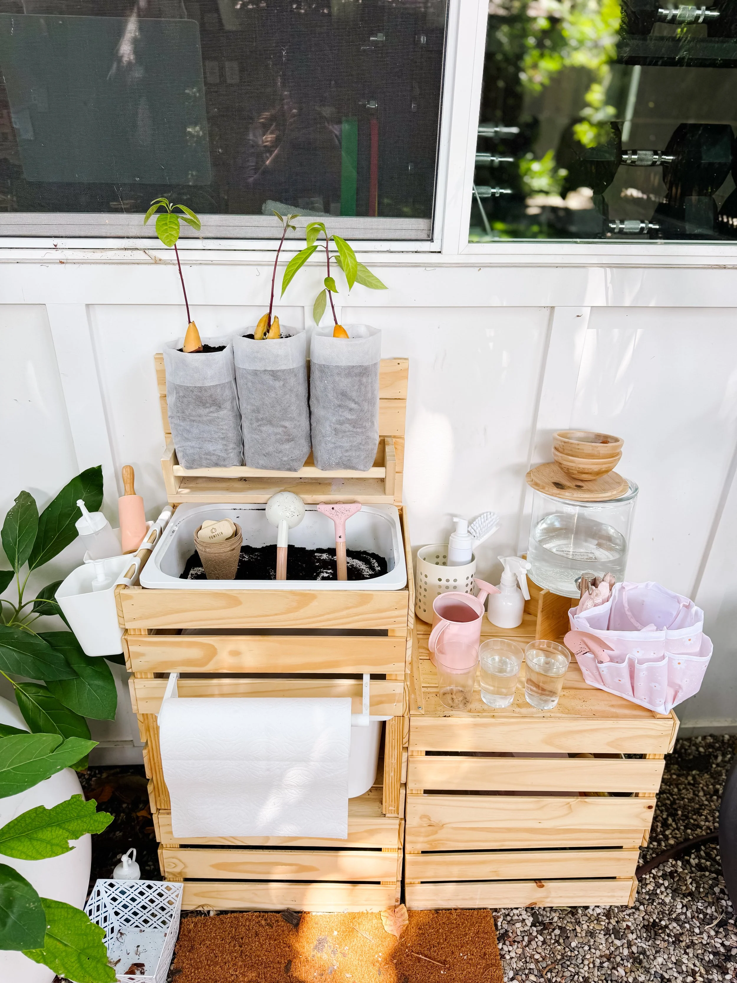 Complete outdoor mud kitchen garden setup with avocado seedlings, grow bags, soil, watering cans and gardening tools.