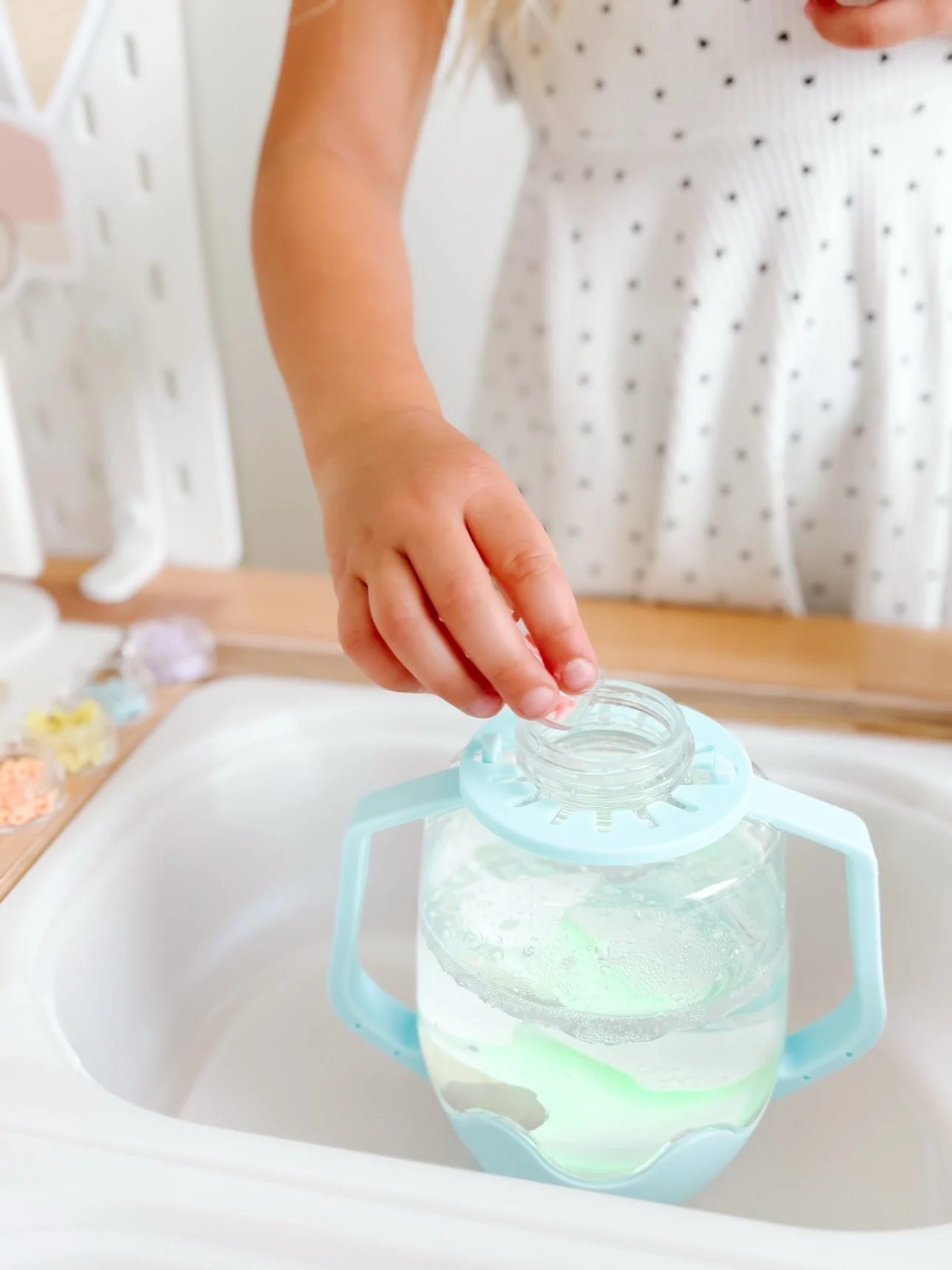 A toddler's hand holding a small water jug over a sensory bin