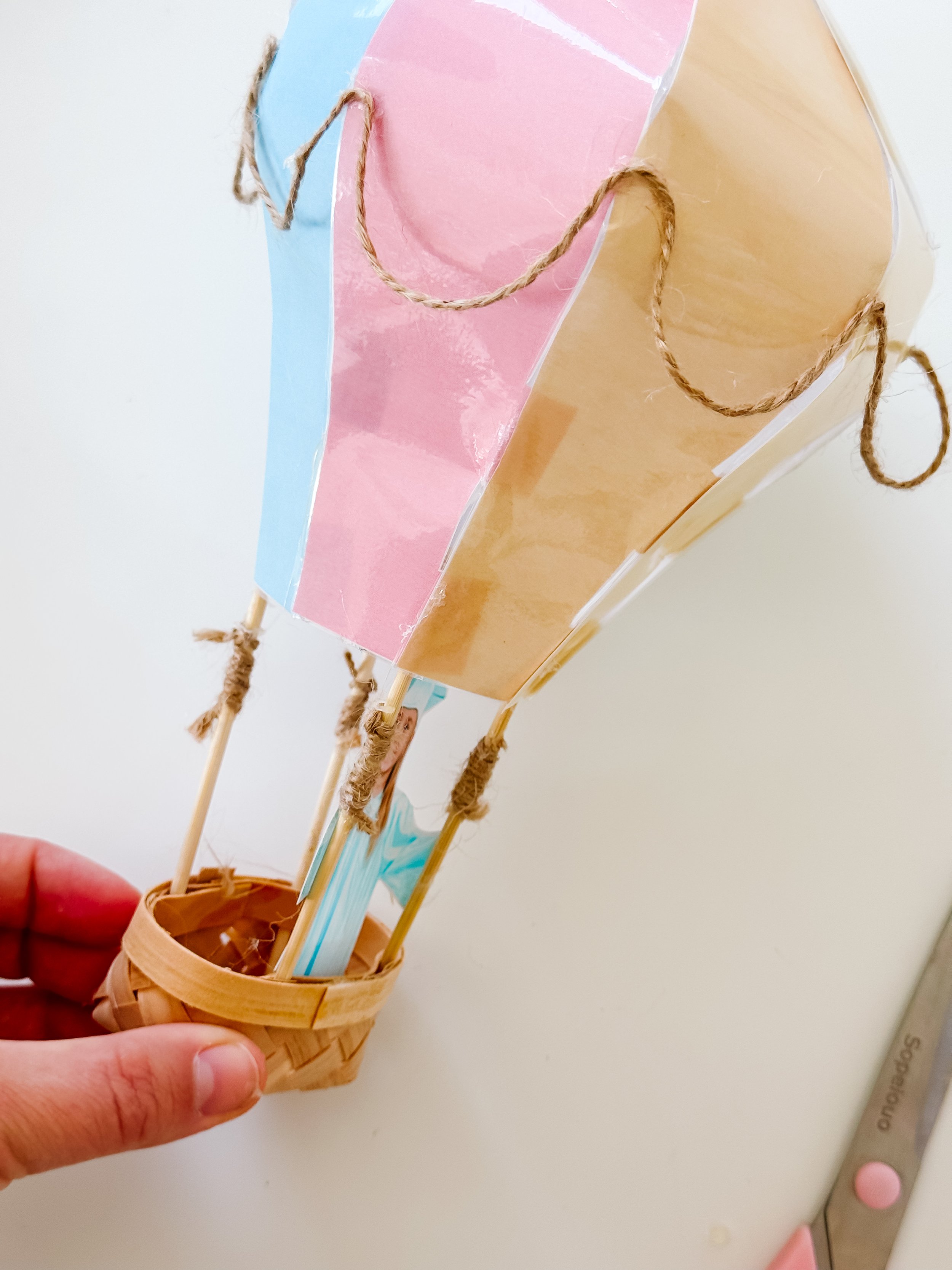 Pastel paper balloon attached to a miniature woven basket using four wooden supports wrapped with twine.