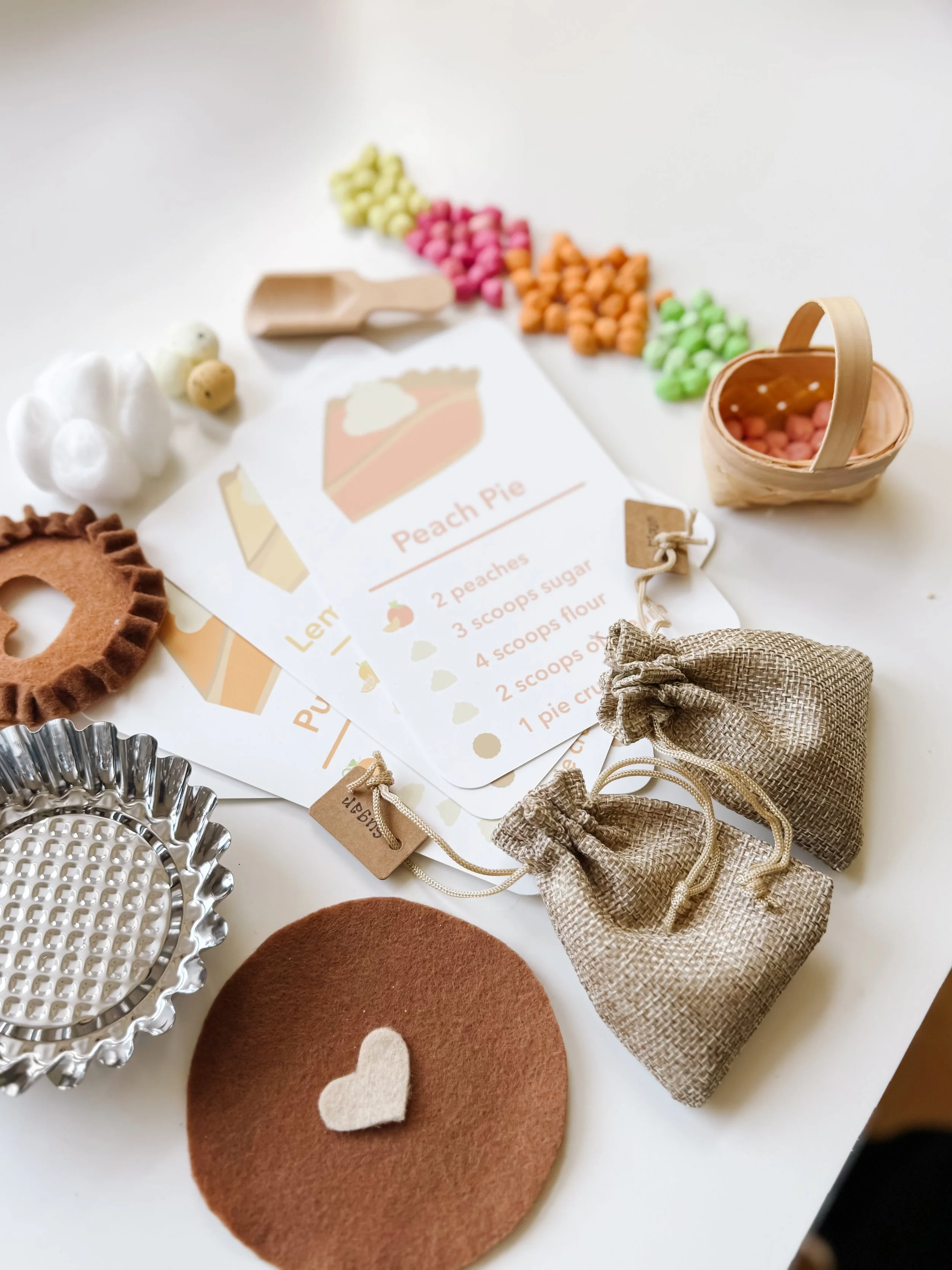 Angled view of the Pie Making Sensory Kit with the Peach Pie recipe card, felt crust topped with a cream felt heart, pie tin, ingredient bags, and chickpea fillings