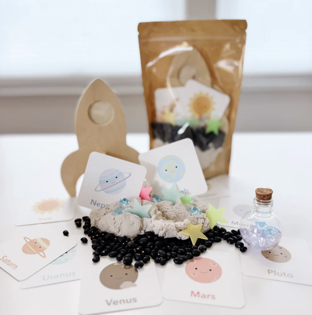 Space themed sensory play kit with moon sand, black beans, glow in the dark stars, planets match game cards and a wooden rocket ship.