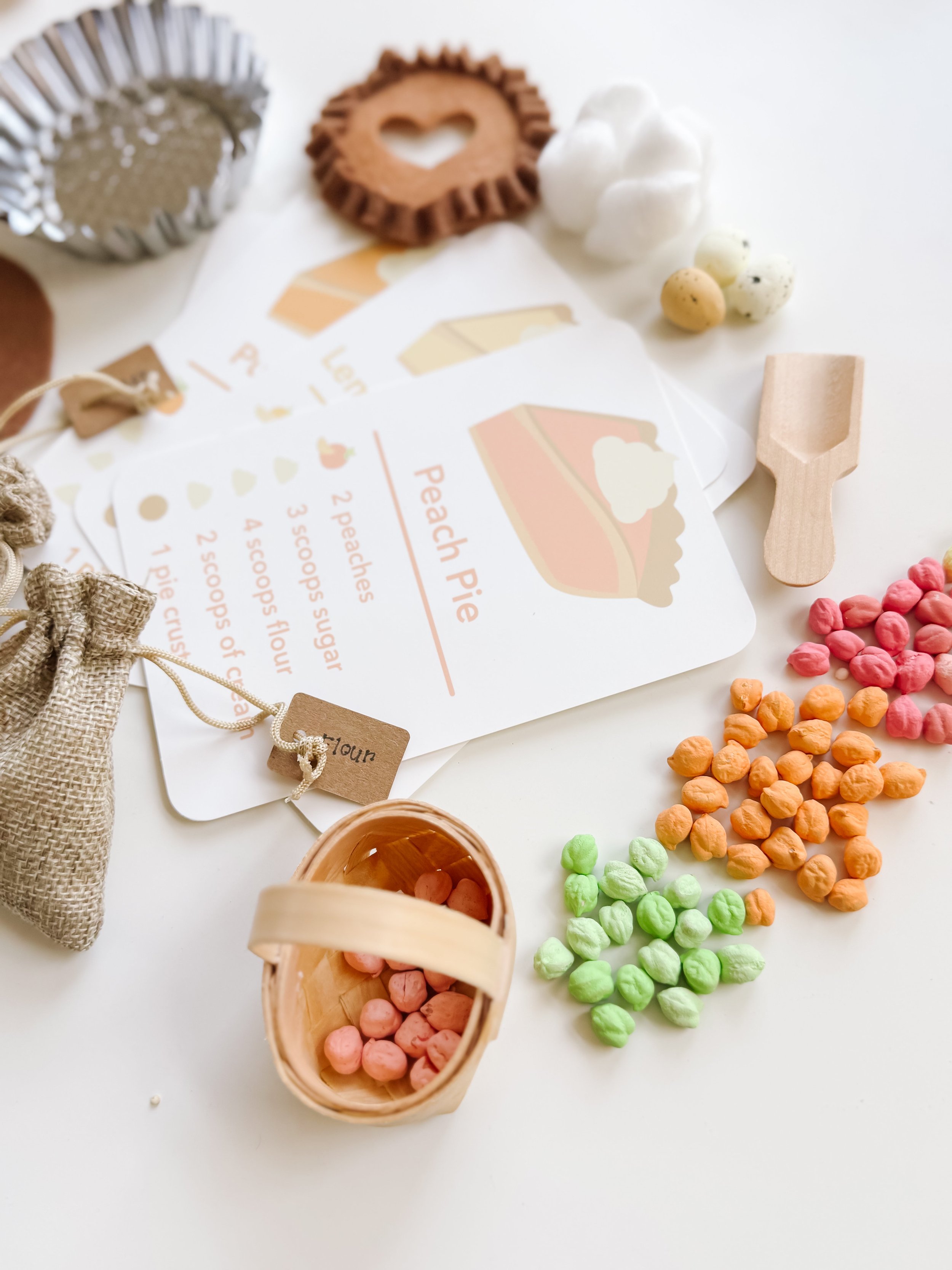 Pie recipe cards fanned out beside a wicker basket of chickpea filling, a wooden scoop, foam eggs, and piles of colored chickpeas