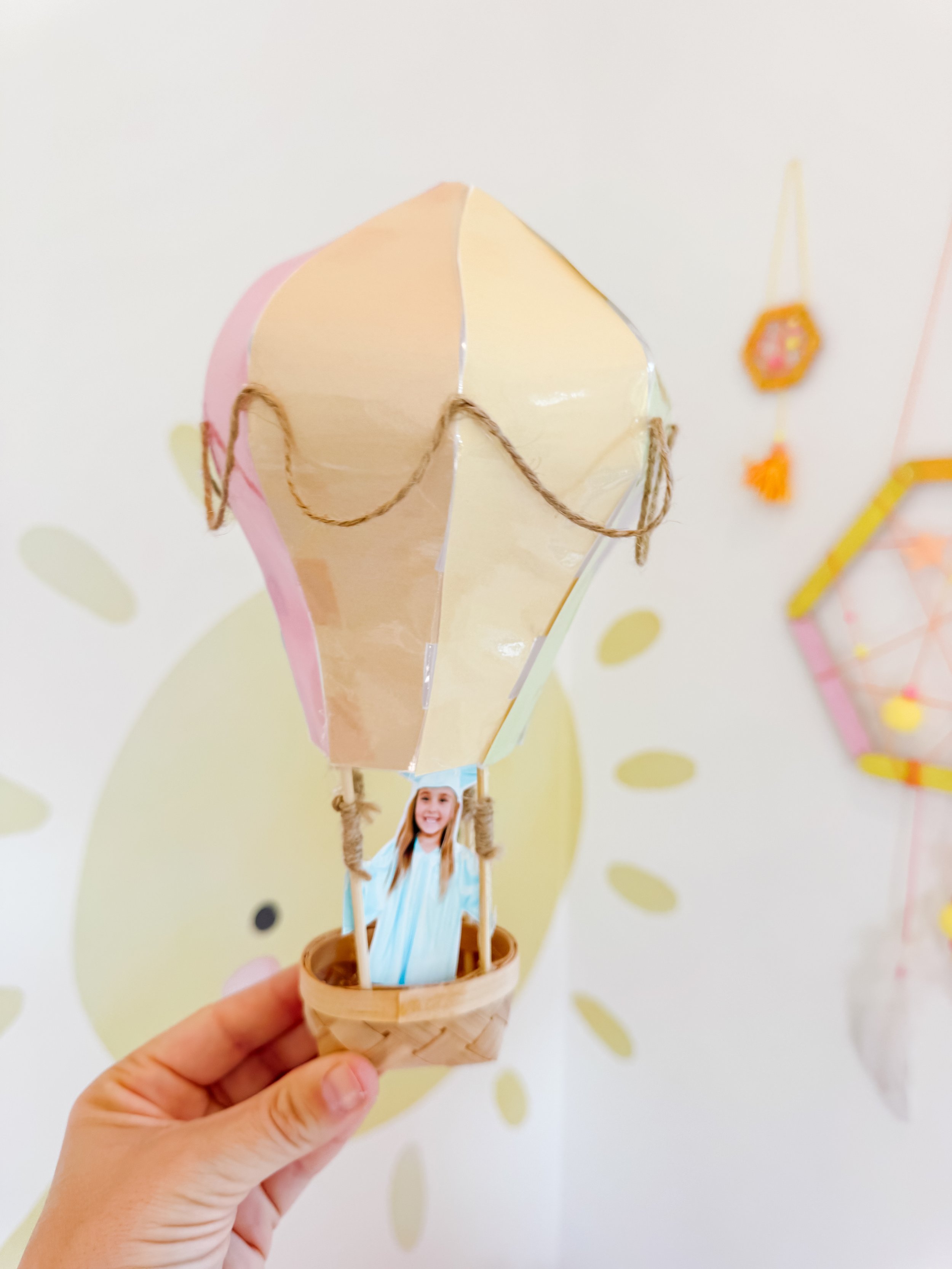 Completed pastel hot air balloon graduation craft held in front of a cheerful sun backdrop.