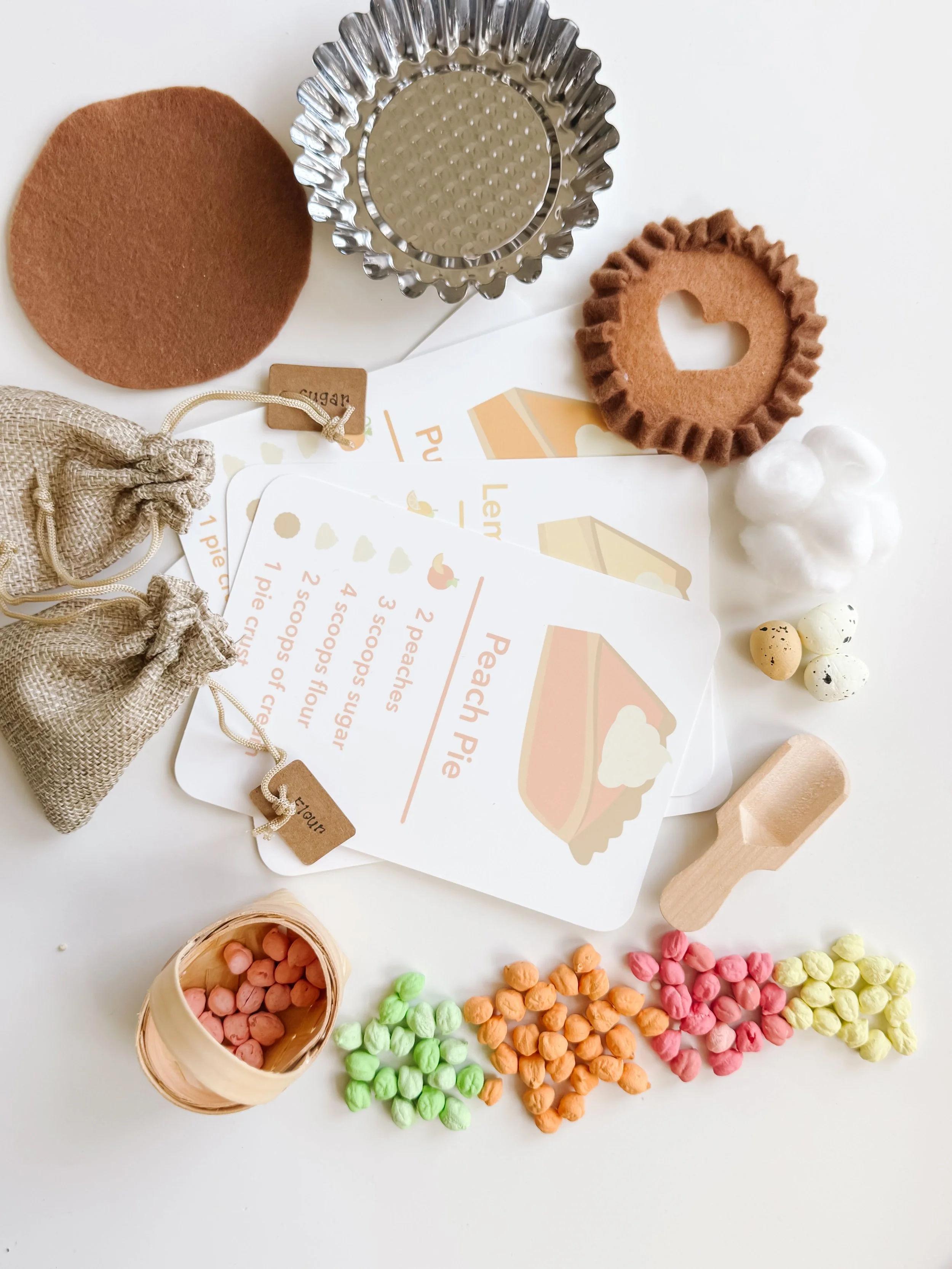 Top-down view of the Pie Making Sensory Kit: metal pie tin, felt pie crust pieces, recipe cards, burlap sugar and flour bags, wooden scoop, foam eggs, and colored chickpeas