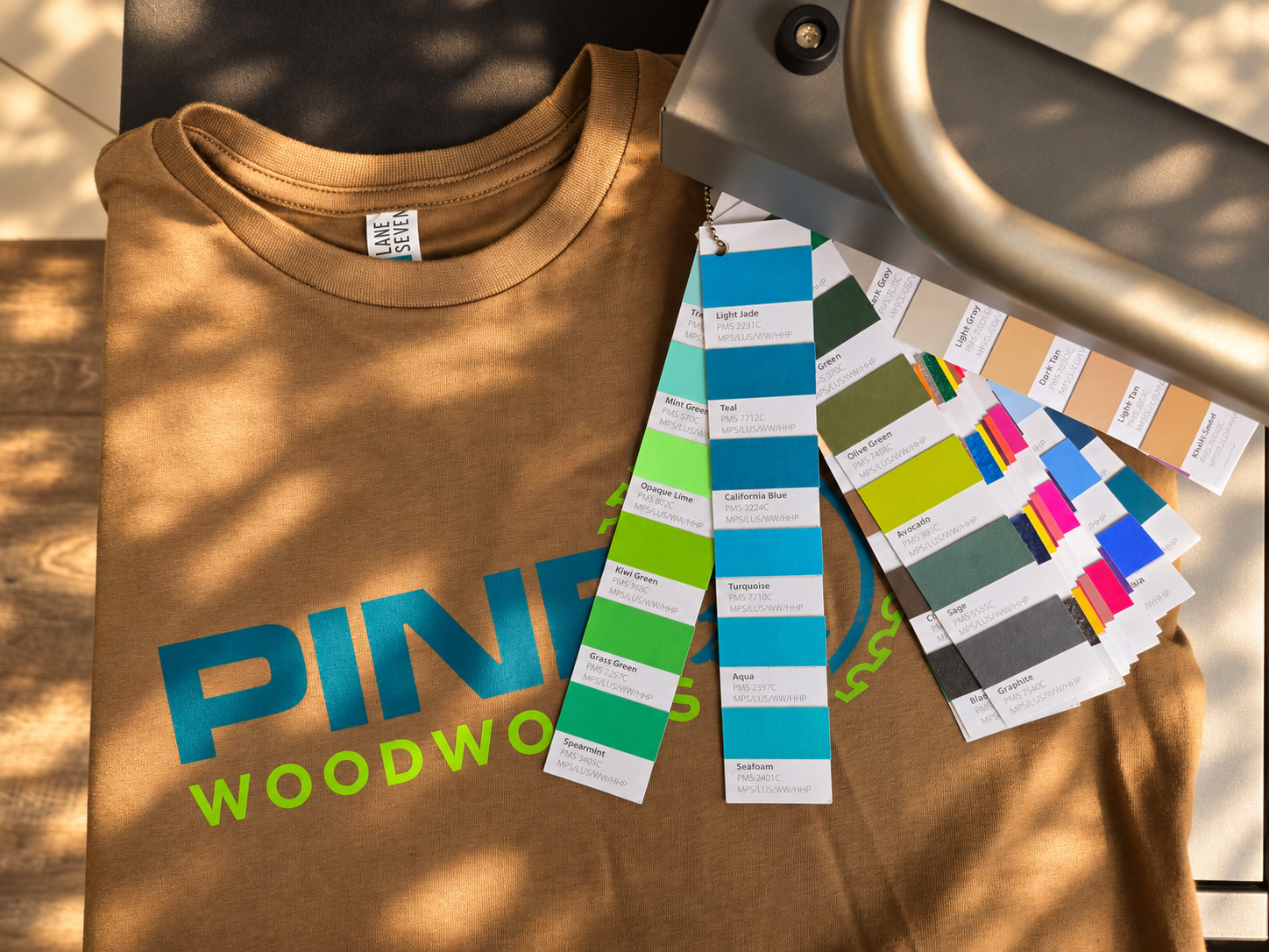 Toasted Coconut Lane Seven garment decorated with Pine Woodworks design alongside Howard Custom Transfers ink color swatch fans and heat press
