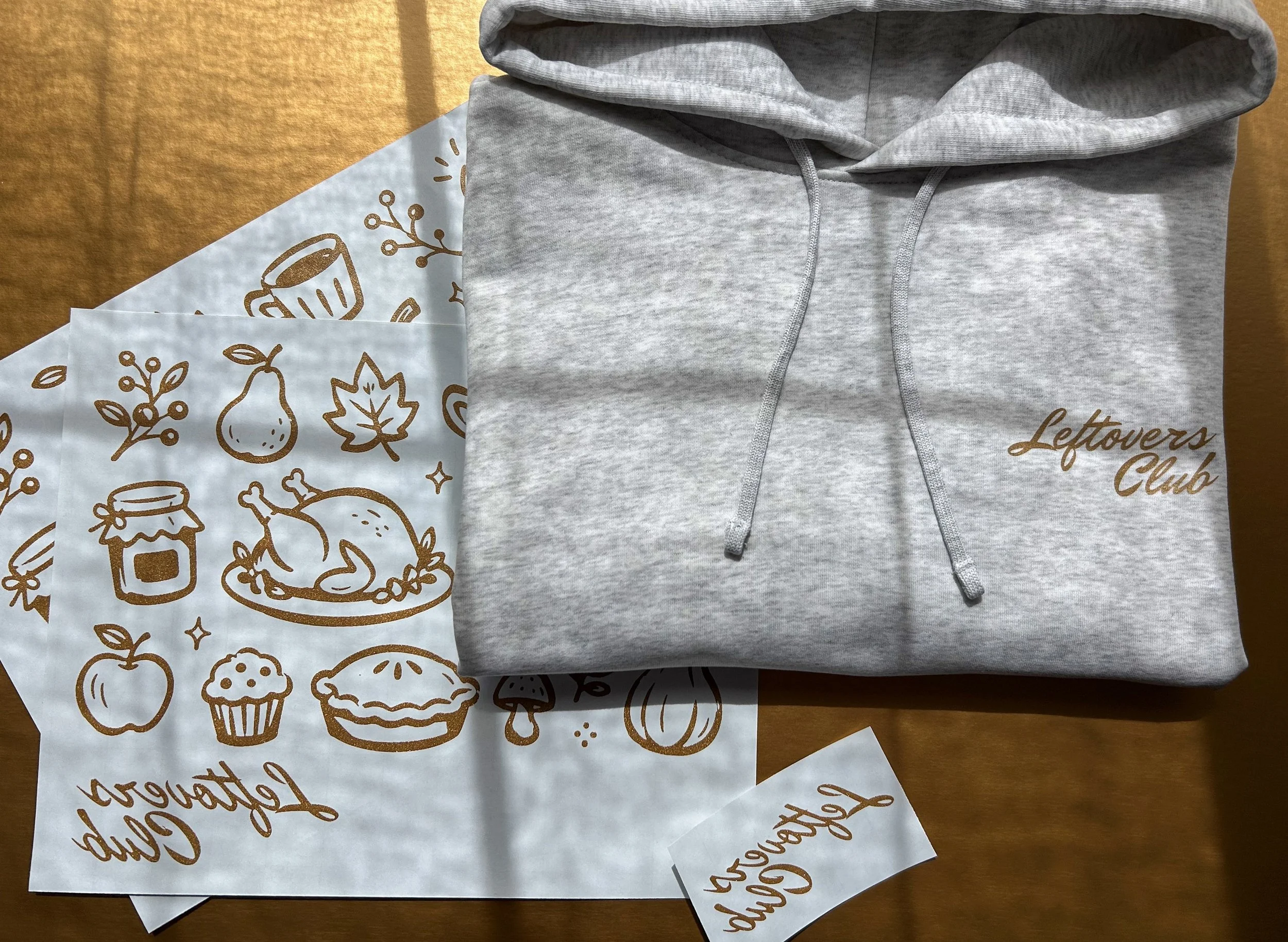 Folded ash grey Stanley/Stella hoodie with a Copper Shimmer Leftovers Club chest print beside matching Howard Hot Peel transfers.