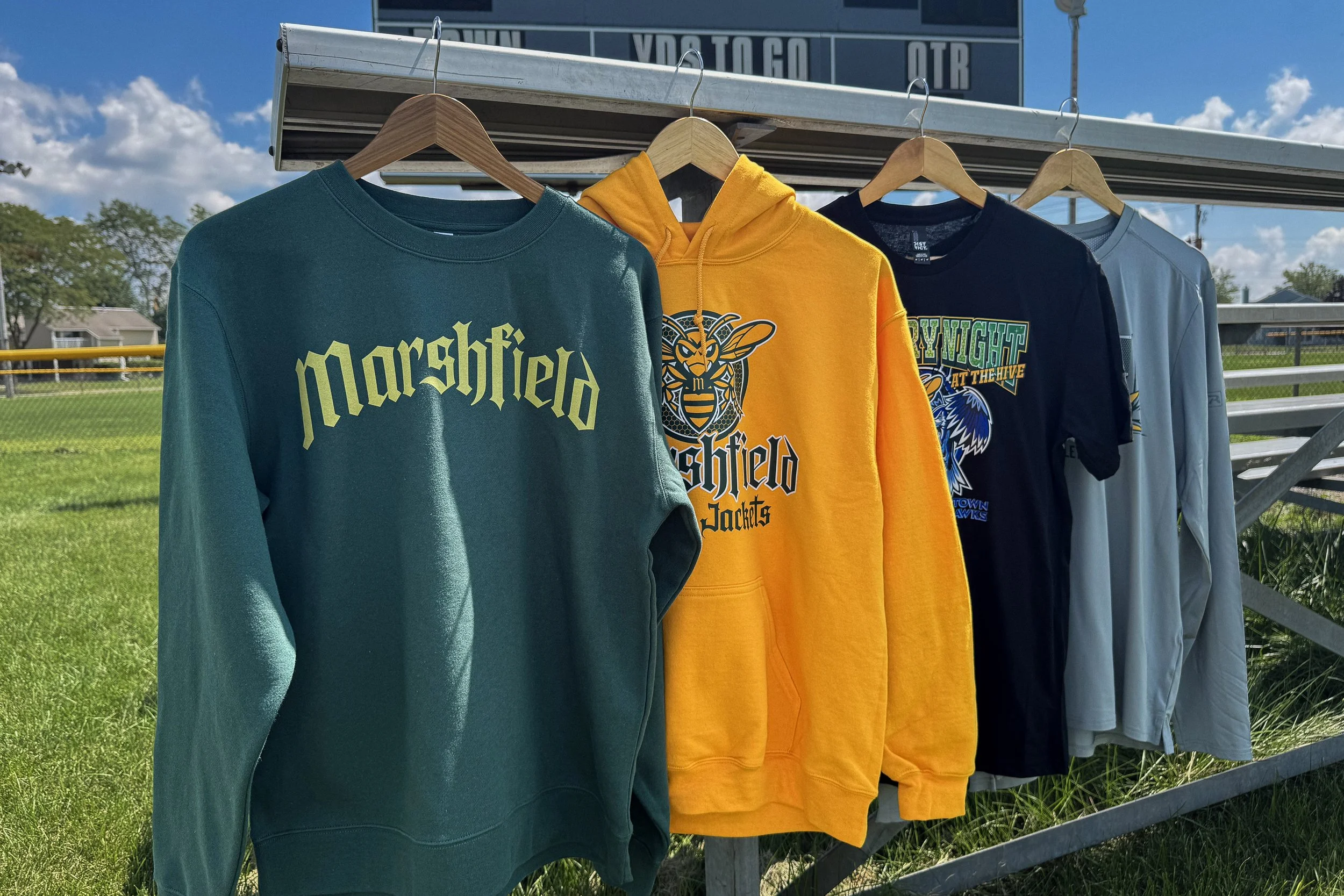 Four-piece Marshfield school spirit wear collection featuring coordinated heat transfer designs in a focused green and gold palette