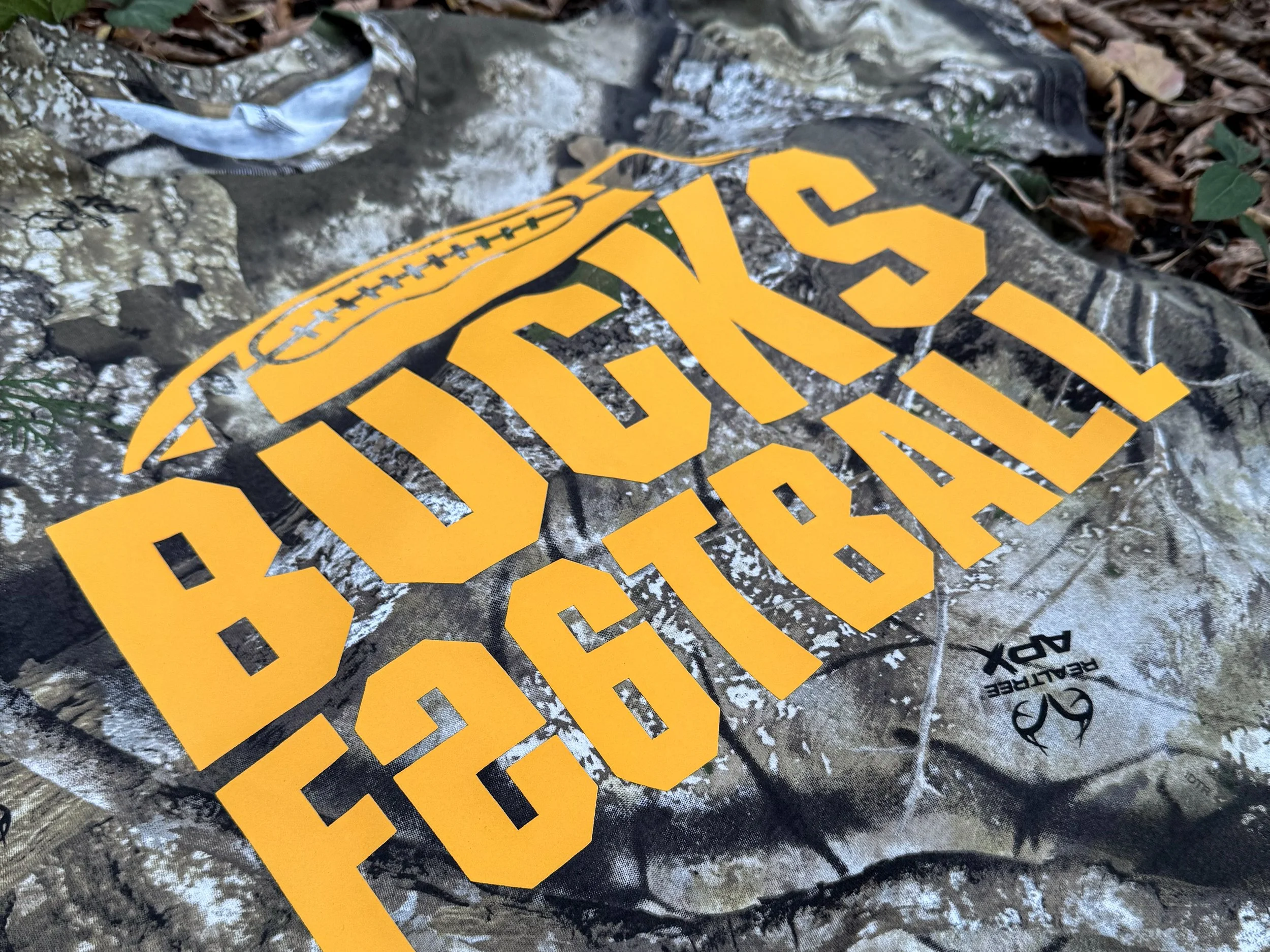 Bold Athletic Gold Bucks Football lettering creates a clear focal point over a busy Realtree APX camo tee.