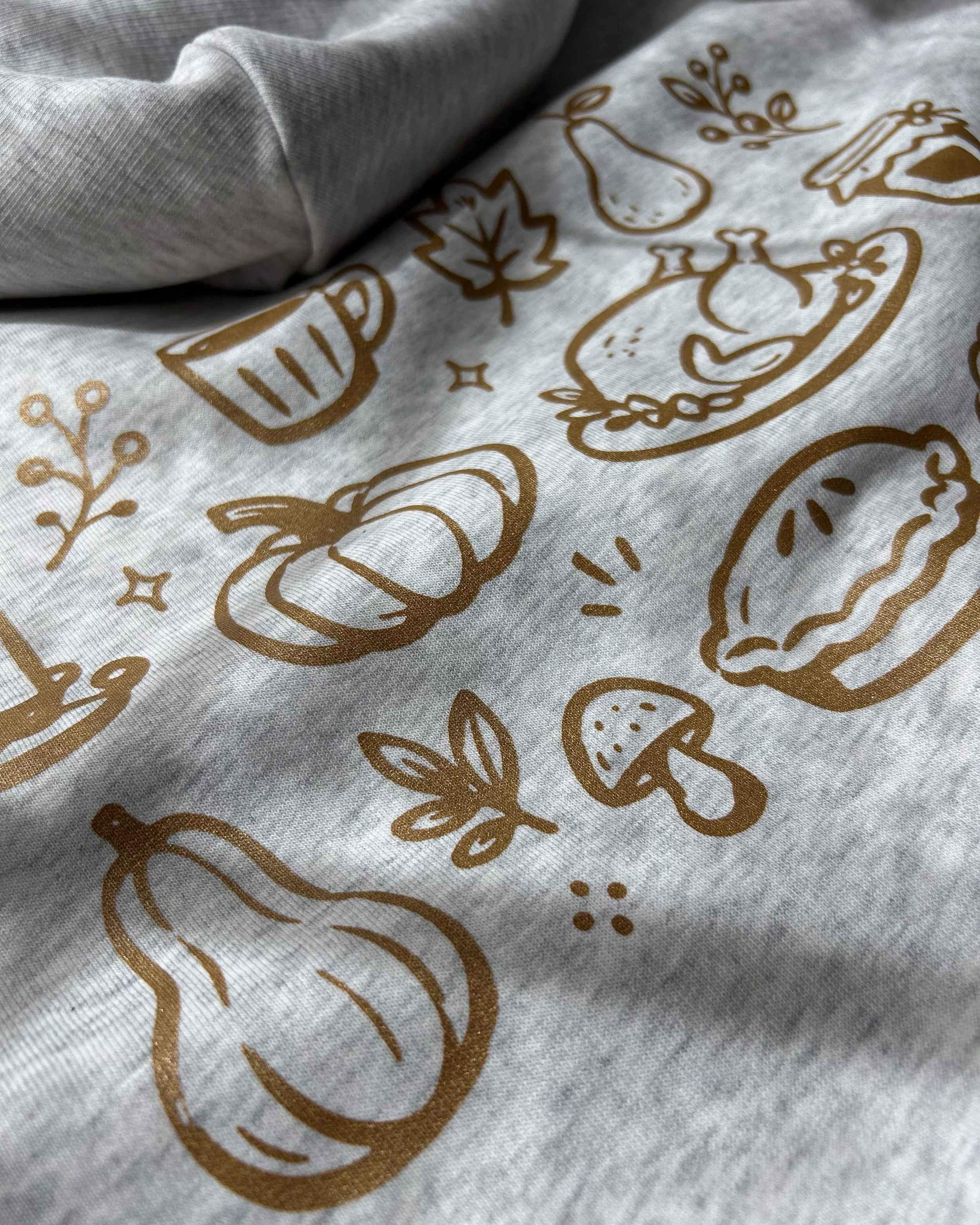 Close-up of illustrated fall food and gathering icons printed in Copper Shimmer on an ash grey Stanley/Stella hoodie.