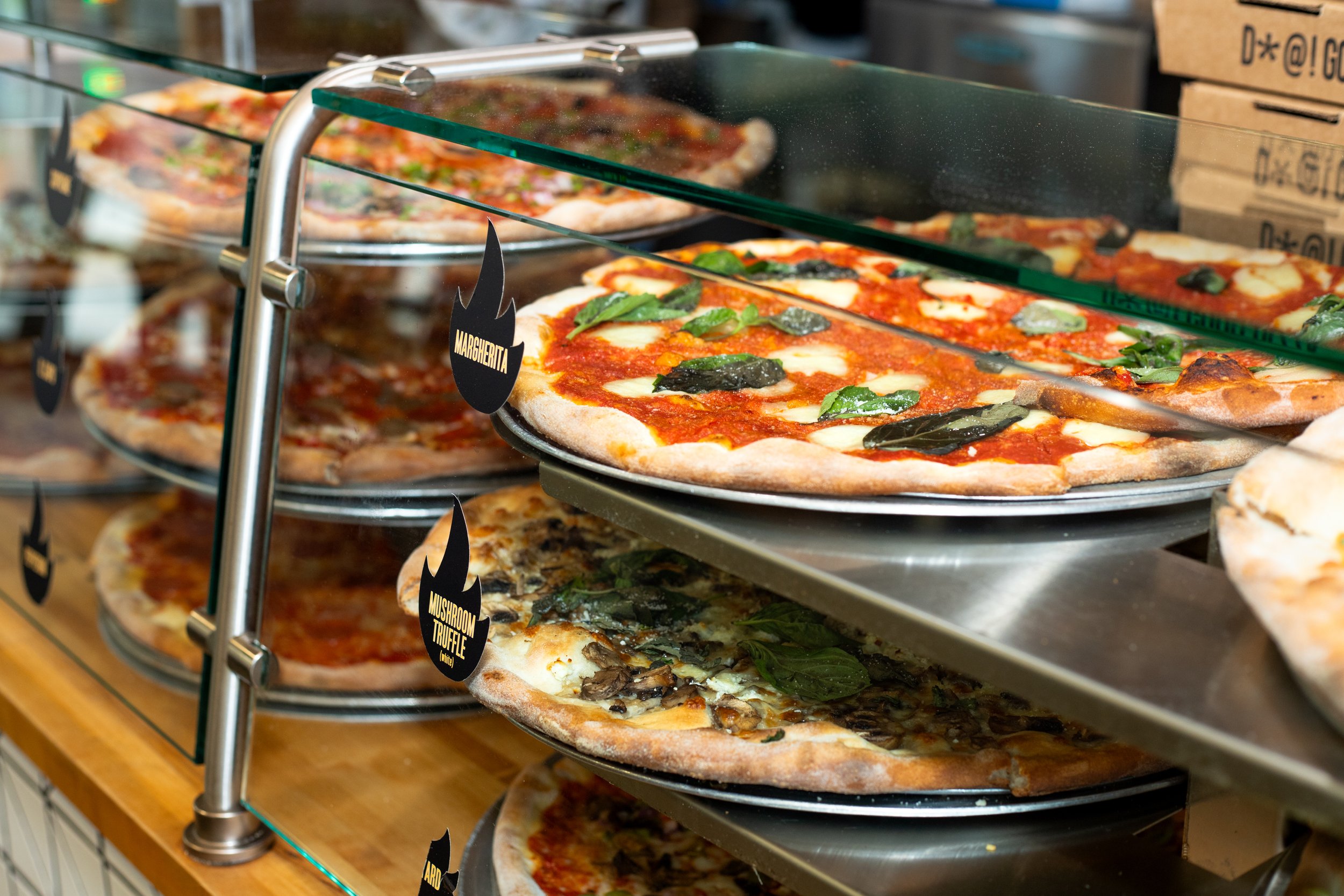 Pizzas on metal trays displayed in a glass case, including a Margherita with tomato sauce and basil, stacked on multiple shelves in a restaurant.
