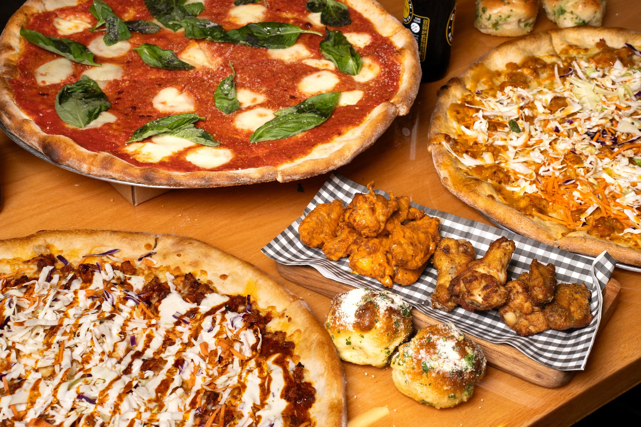 Assorted pizzas with cheese, vegetables, and meats, along with fried chicken and dinner rolls on a wooden table.