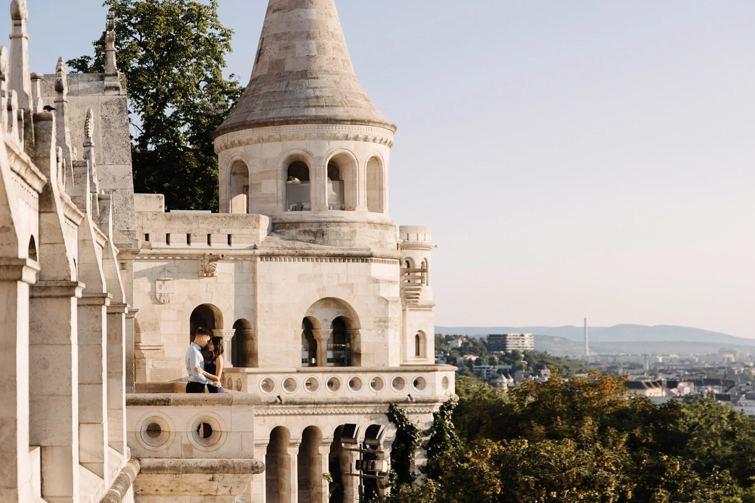 A couple standing close together on a balcony of a historic white stone building with intricate architectural details, overlooking a cityscape with trees and modern buildings in the distance during late afternoon or early evening.