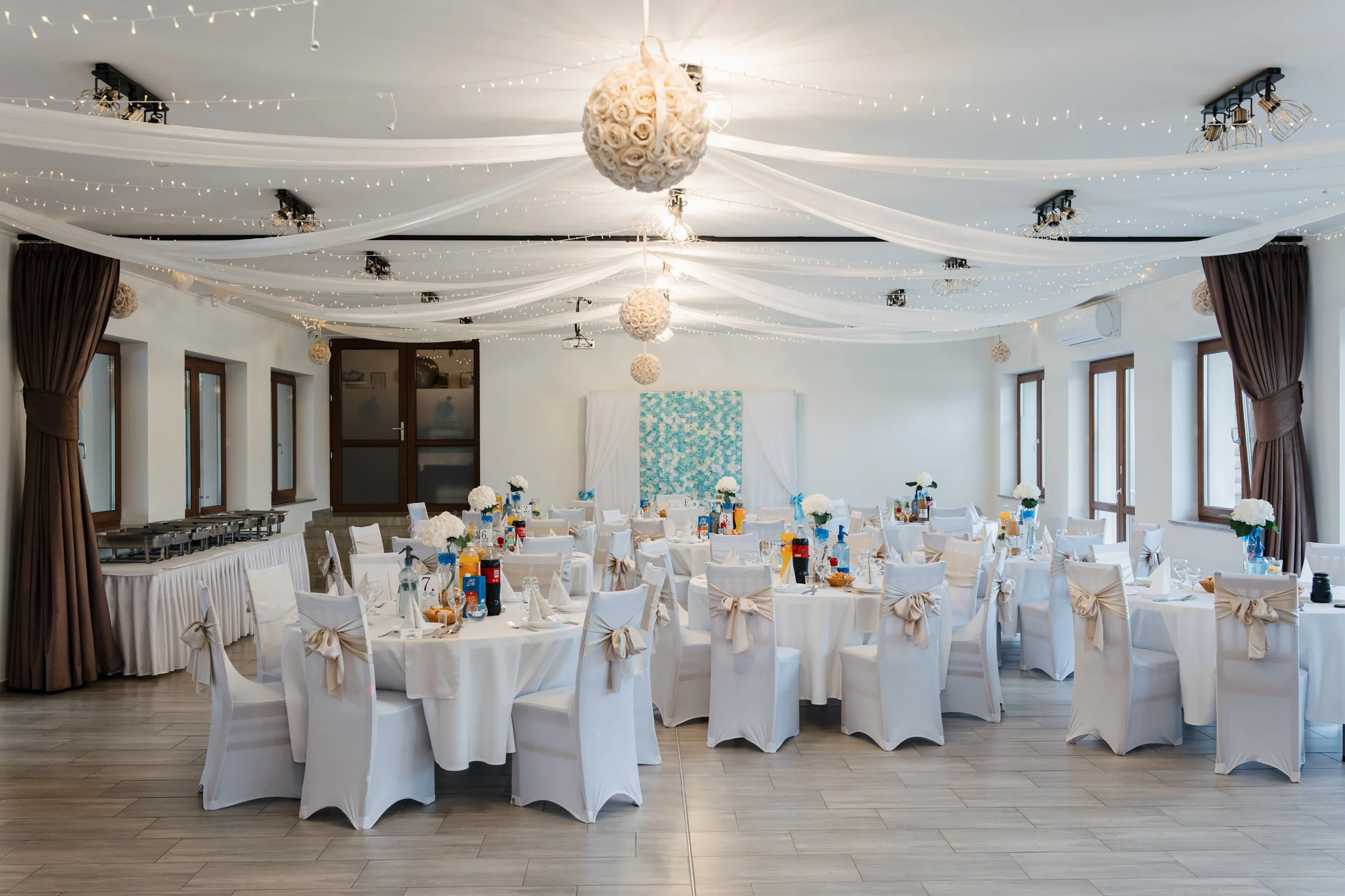 Banquet hall decorated for a celebration with white tablecloths, chairs with bows, and floral centerpieces. The ceiling has white drapes, string lights, and hanging floral arrangements. There are windows along the side walls with brown curtains.