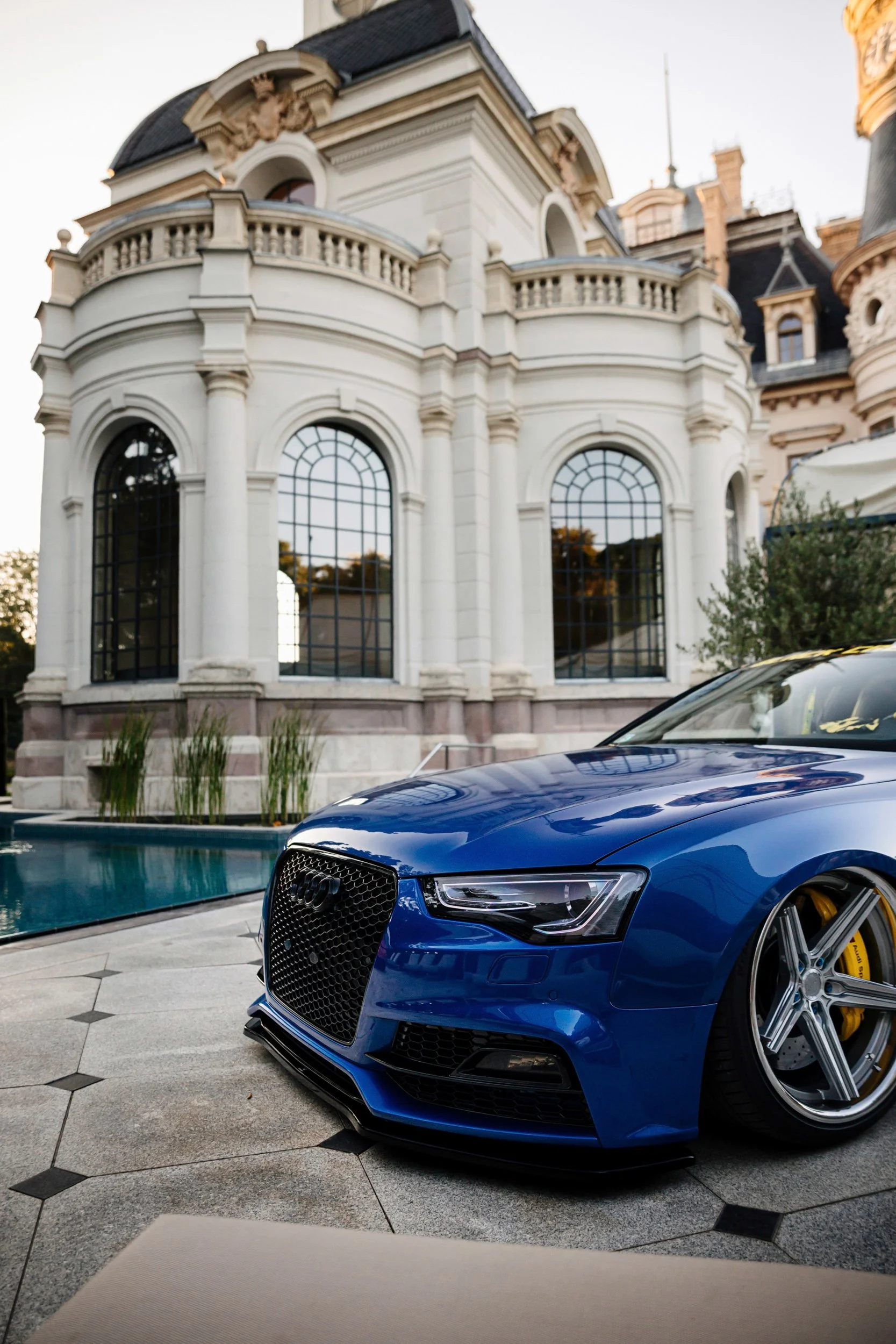 A shiny blue Audi sports car parked in front of a luxurious mansion with a pool and ornate architecture.