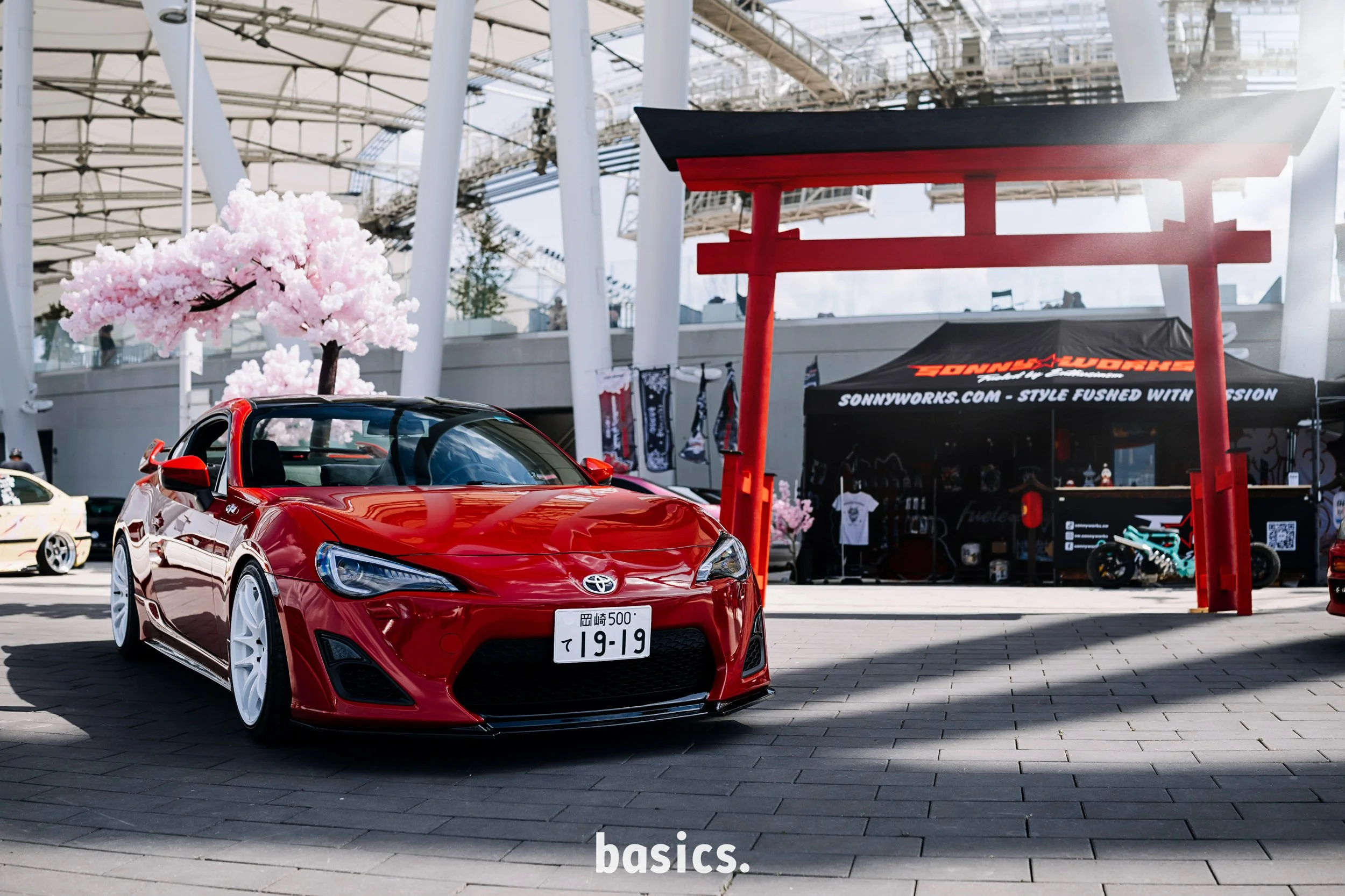 A red sports car with white wheels and Japanese license plate parked in front of a cherry blossom tree and a Japanese torii gate at an outdoor event space.