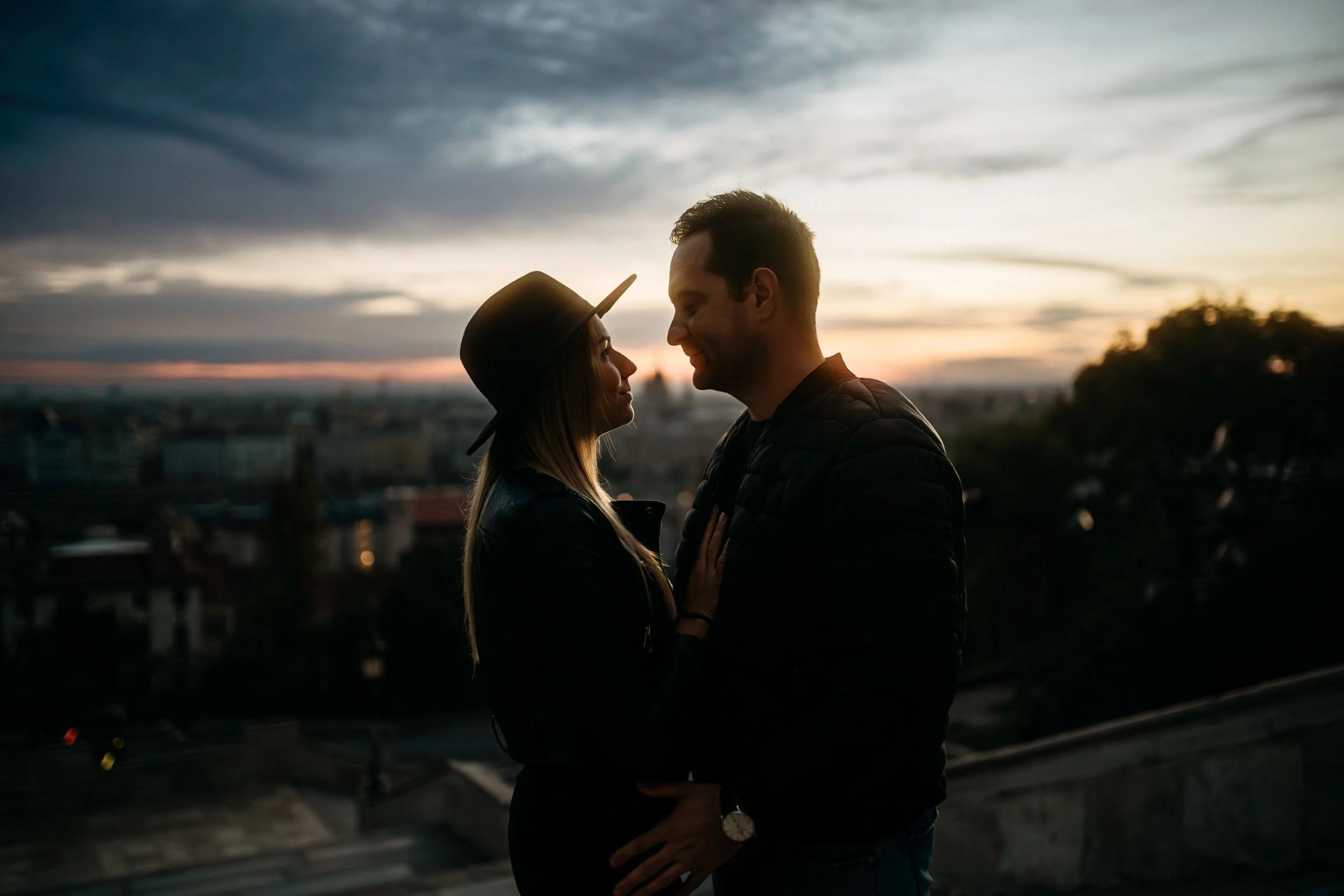 A couple standing close together outdoors at sunset, facing each other with their foreheads nearly touching, smiling softly. The woman is wearing a hat, and both are dressed in dark jackets, with a cityscape blurred in the background under a cloudy s