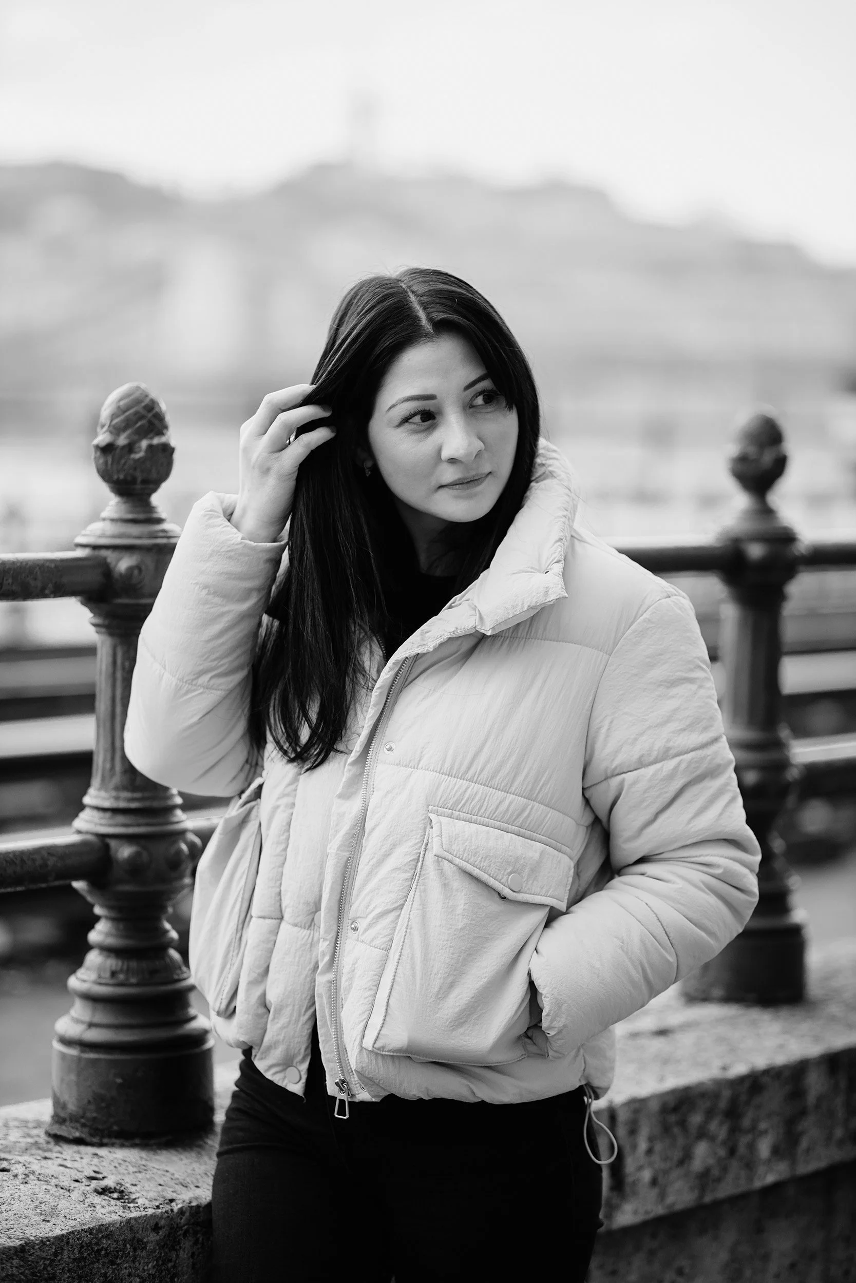 Black and white photo of a young woman with long dark hair, wearing a light-colored quilted jacket, standing near a decorative metal railing outdoors. She is touching her hair with one hand and looking to her right.