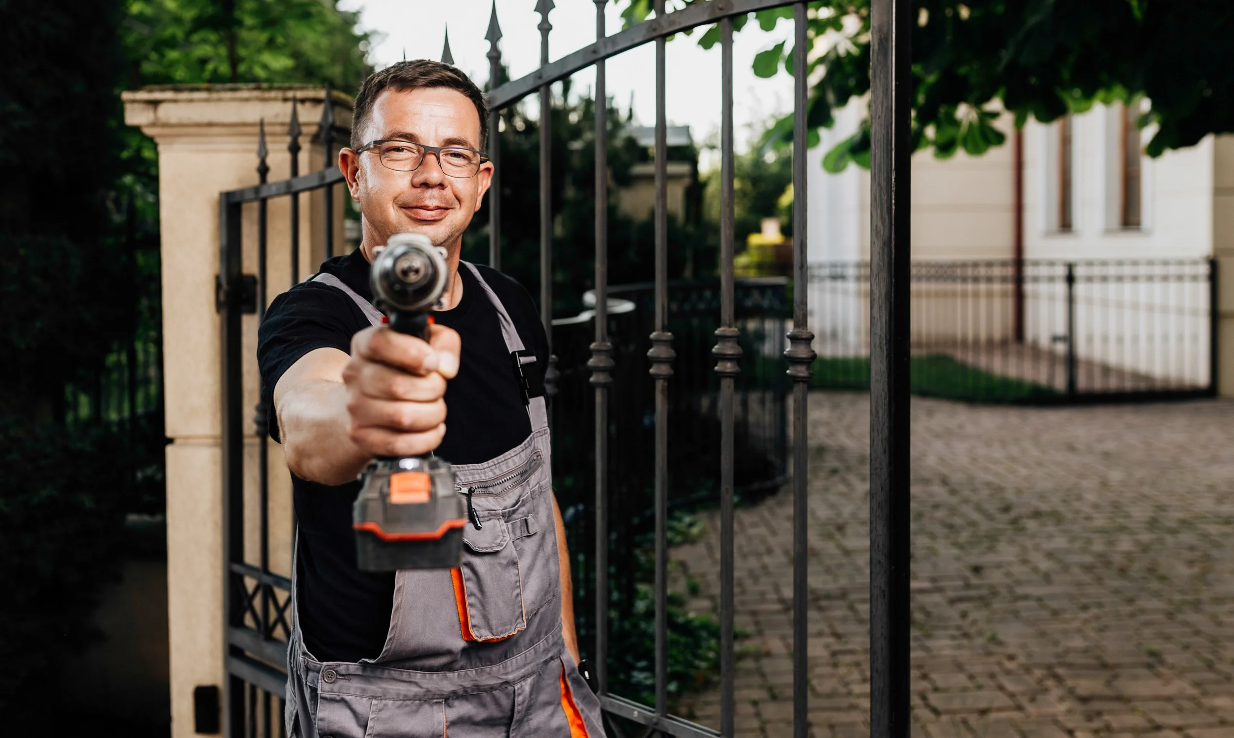 A man with glasses and a short hairstyle, wearing overalls and a black t-shirt, holding a power drill pointed towards the camera, standing in front of a black metal gate with stone pillars, greenery, and a house in the background.