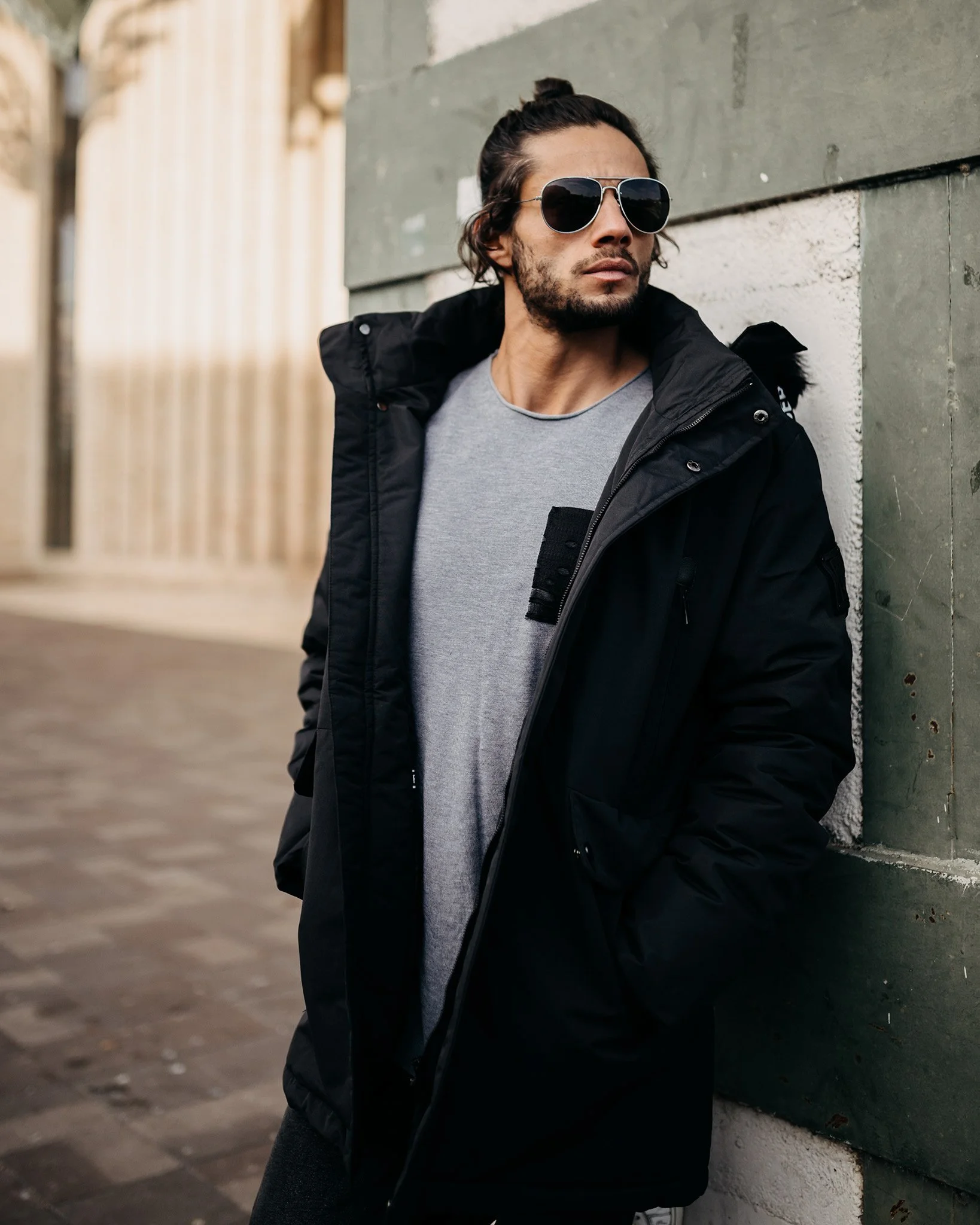 A man with dark hair tied in a top knot, beard, and sunglasses leaning against a green textured wall outdoors. He is wearing a black jacket over a gray shirt.