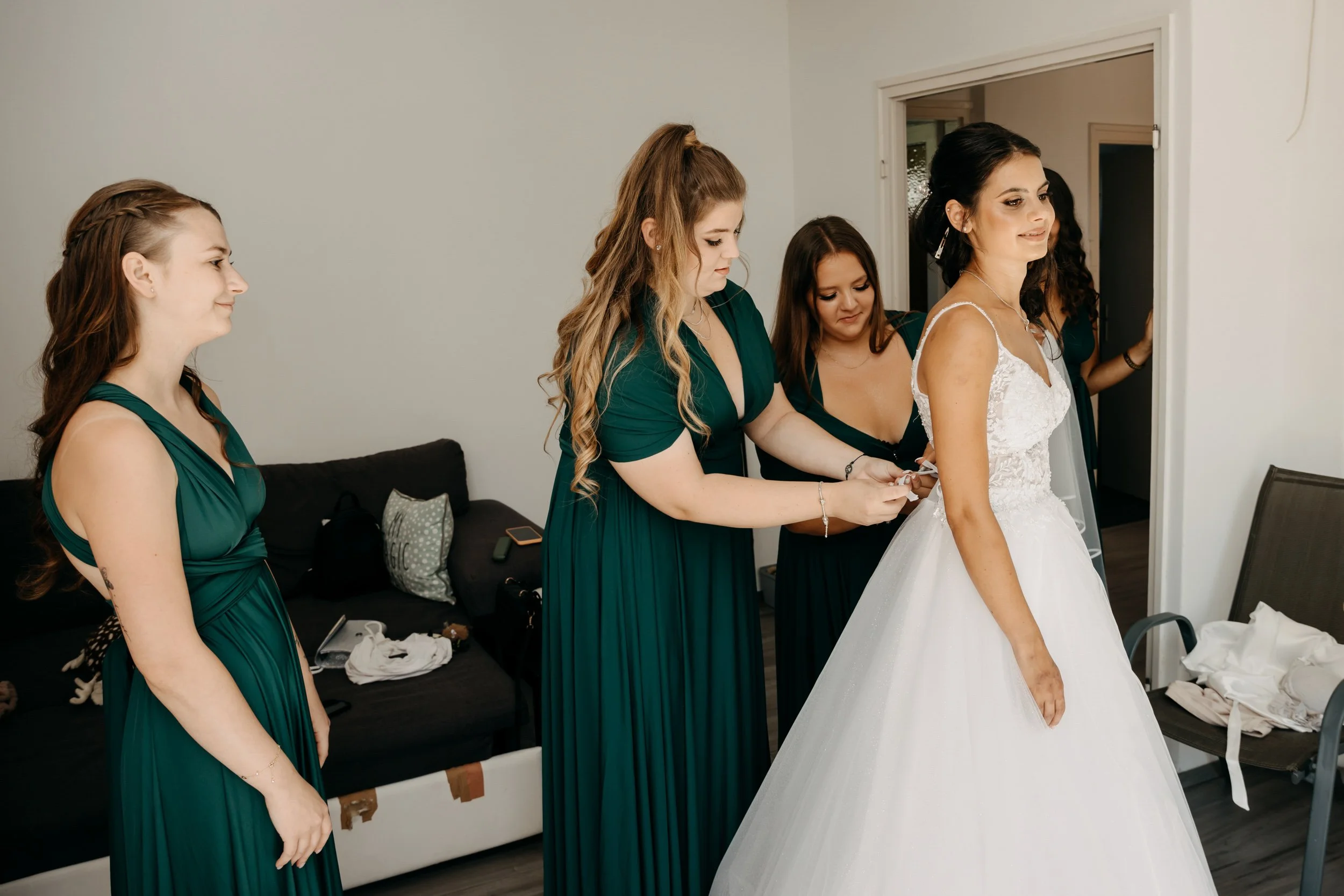 A bride in a white wedding dress standing in front of four bridesmaids dressed in dark green gowns. The bridesmaids are helping the bride with her dress, with one adjusting the back. The room has a black sofa with pillows, and various items are scatt