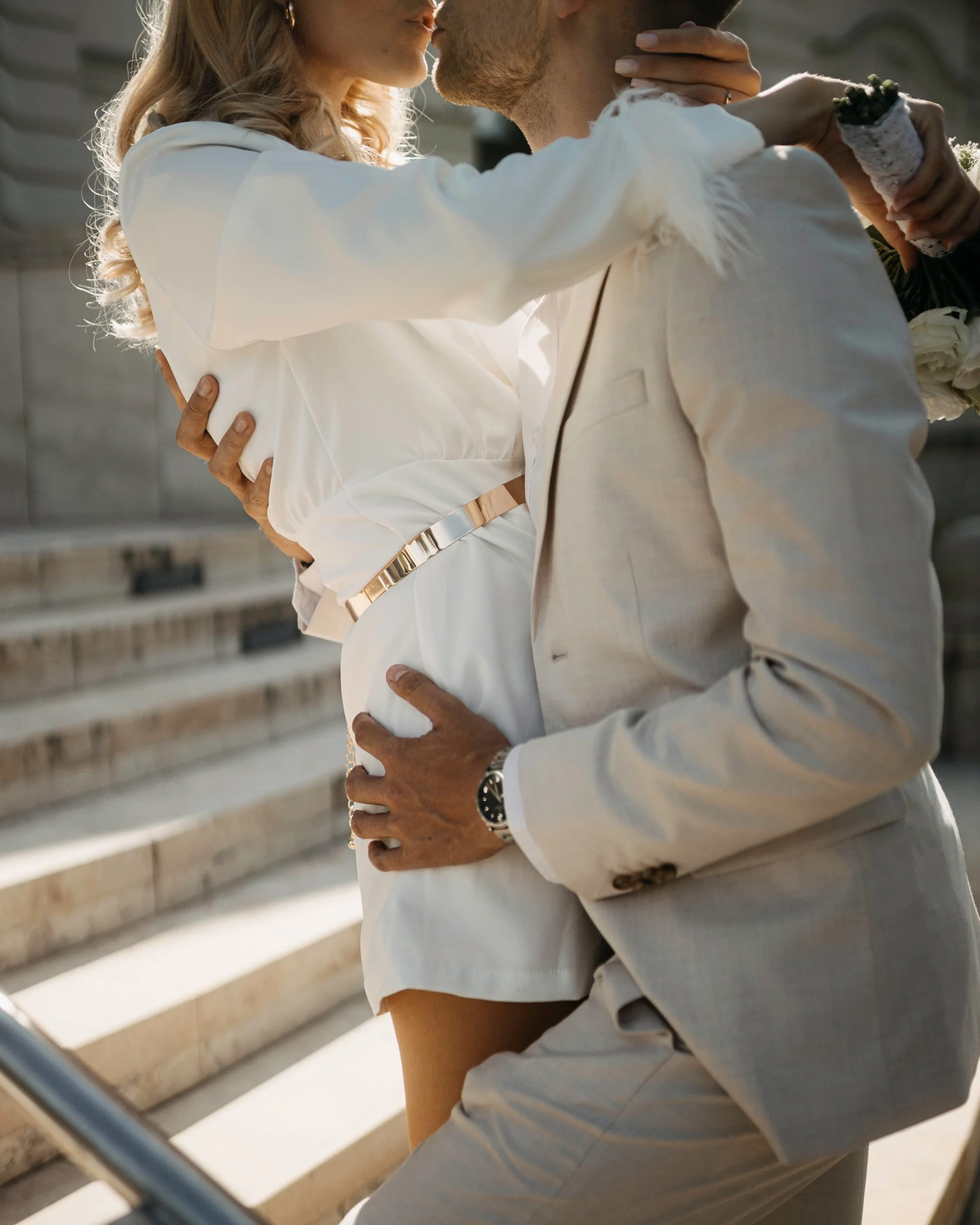 A couple sharing a kiss outdoors, with the woman wearing a white blouse and the man in a light-colored suit, framed by horizontal wooden rails.