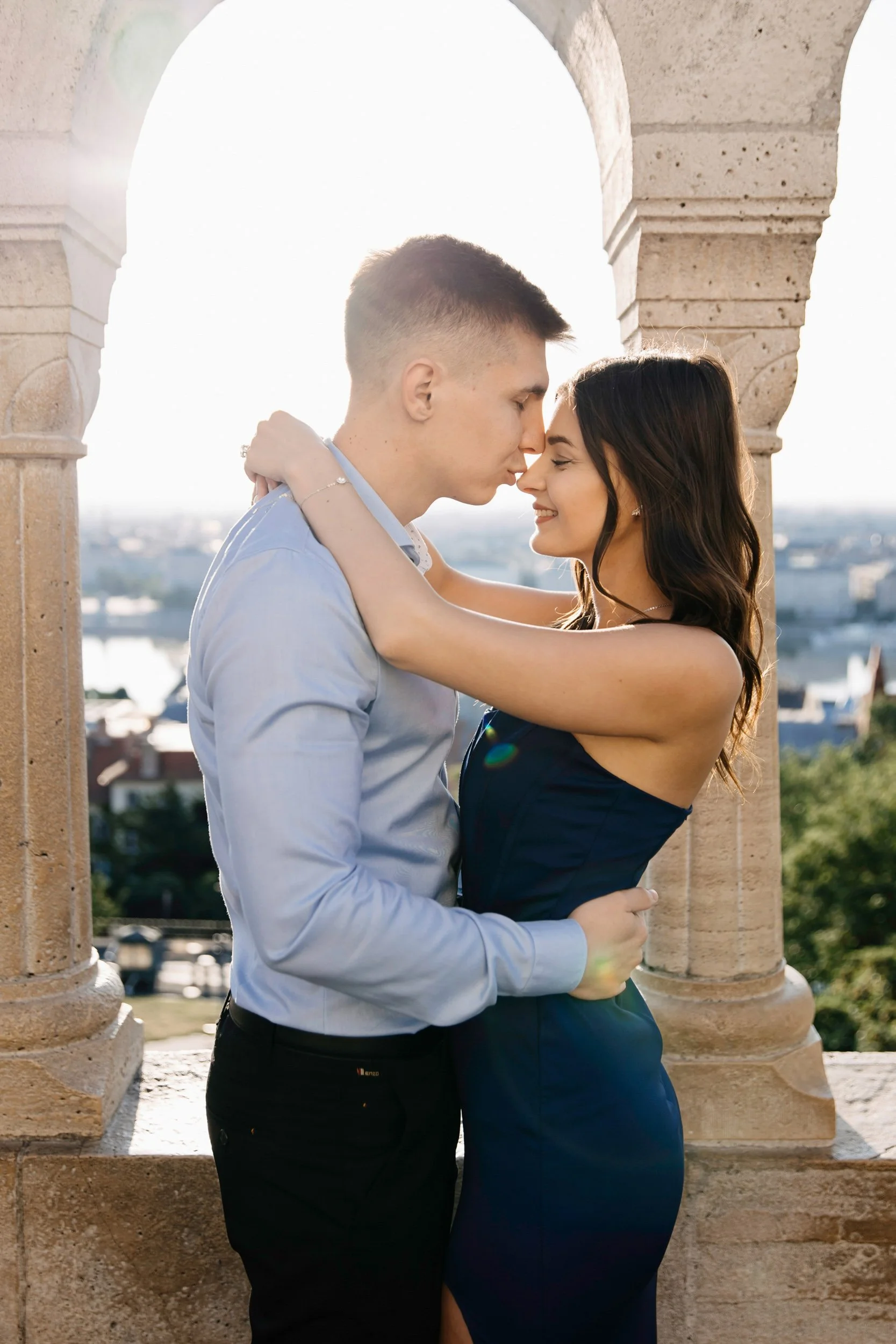 A young couple standing close, with foreheads touching, smiling and embracing, on a balcony with stone columns, overlooking a cityscape.