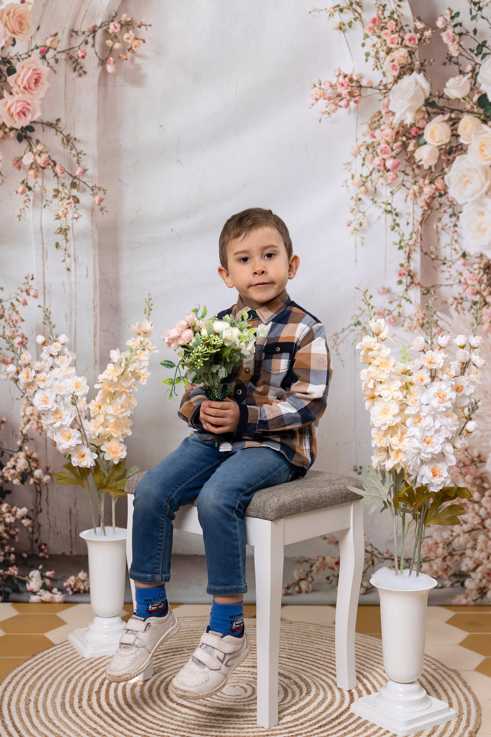 A young boy sitting on a white bench holding a bouquet of flowers, surrounded by pink and white floral decorations and vases with flowers, in an indoor setting with a beige and white patterned rug.