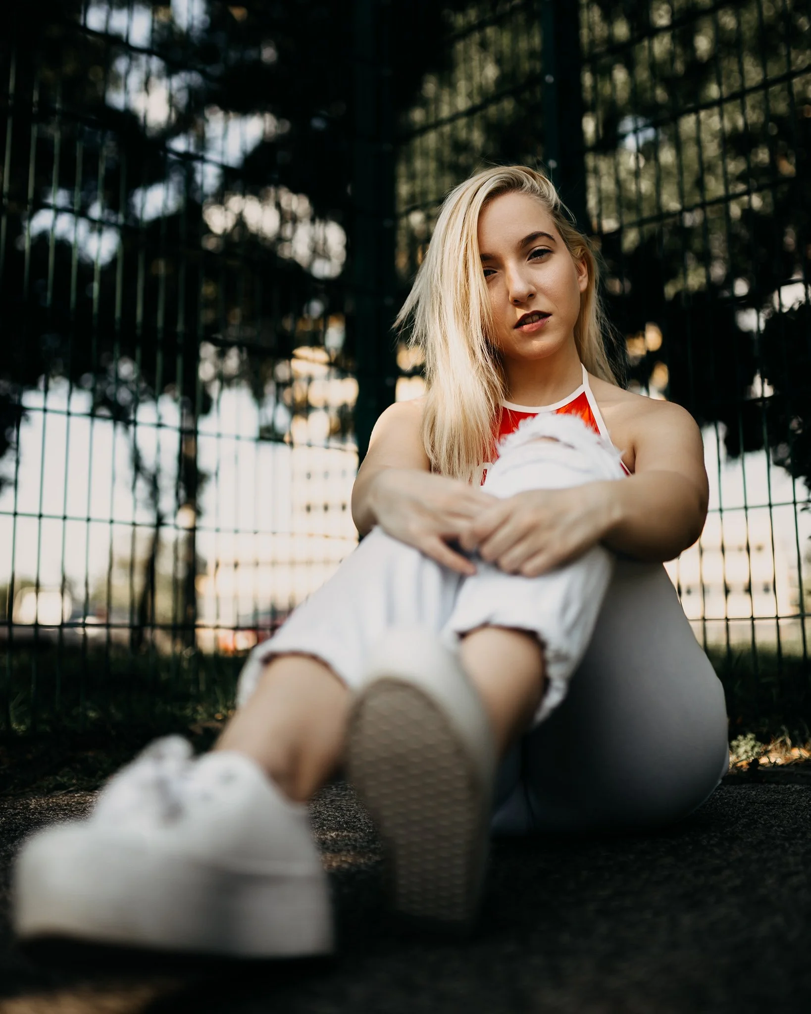A young woman with long blonde hair sitting on the ground near a metal fence outdoors during sunset, looking into the camera with a neutral expression, wearing a red and white top and white pants.