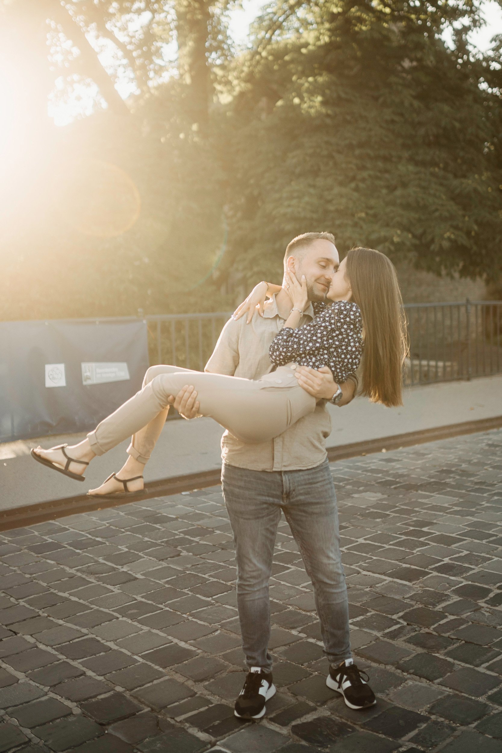 A man is holding a woman in his arms outdoors during the golden hour, with sunlight creating a backlit effect. They are exchanging a kiss, surrounded by trees and a black fence, on a cobblestone street.