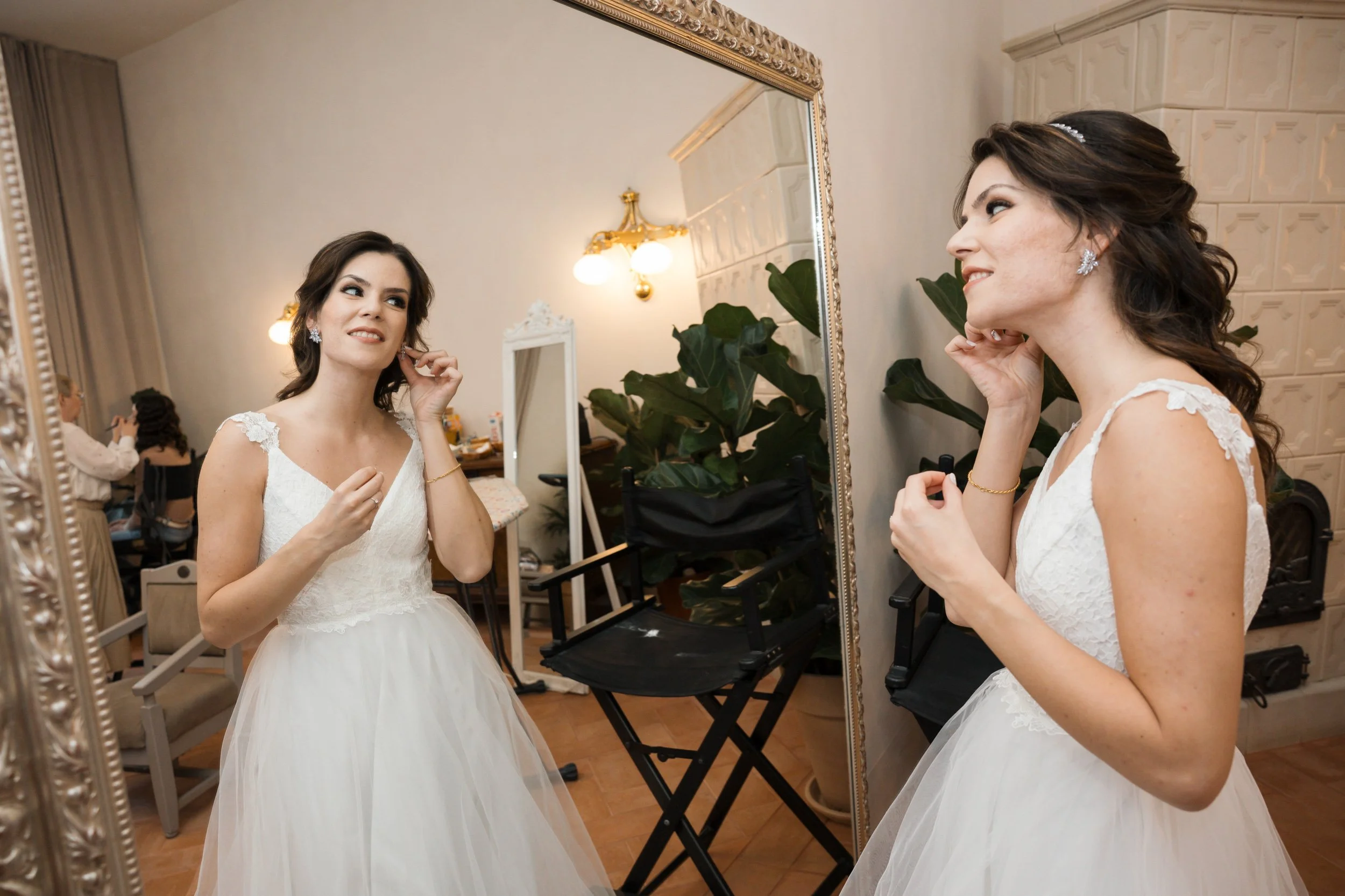 A woman in a white dress is looking at herself in a large mirror while adjusting her earrings. She has dark, wavy hair and is smiling. The room has bright lighting, with a plant and some makeup items visible in the background.
