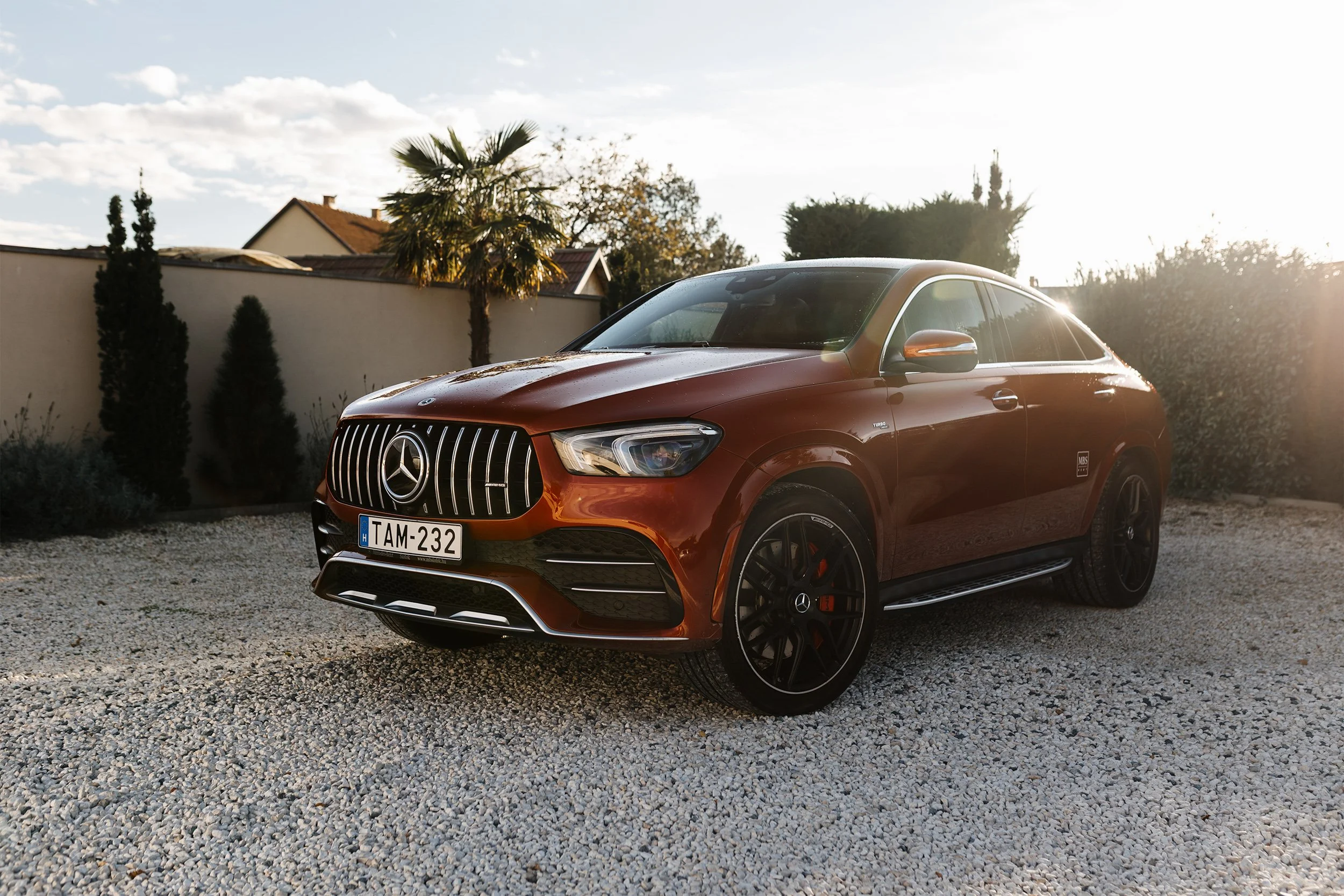 A red Mercedes-Benz SUV parked on a gravel driveway, with buildings and trees in the background, during sunset.