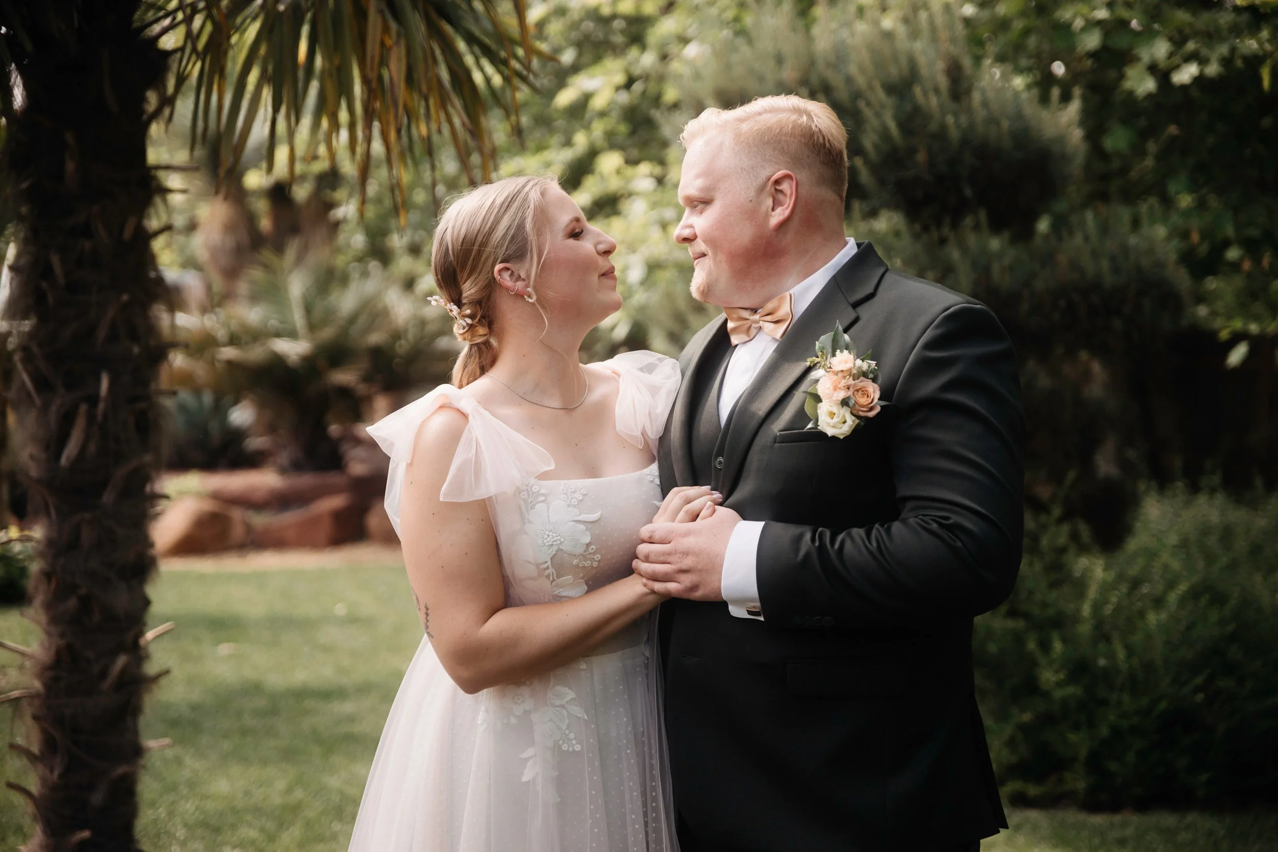 A bride and groom standing close together outdoors, holding hands, looking into each other's eyes, with greenery in the background.