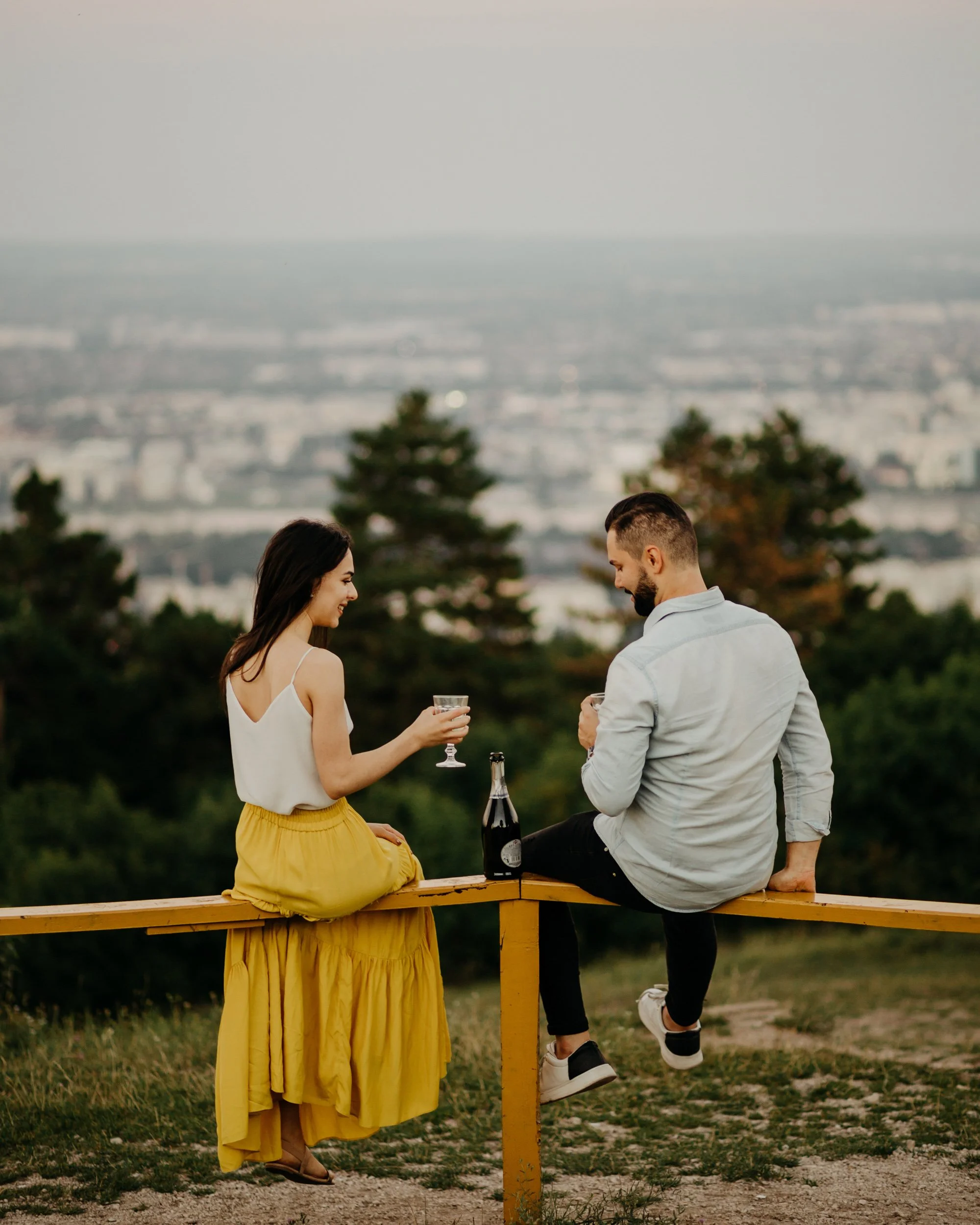 A man and a woman sitting on a wooden bench outdoors, holding drinks, with a cityscape and trees in the background during sunset.