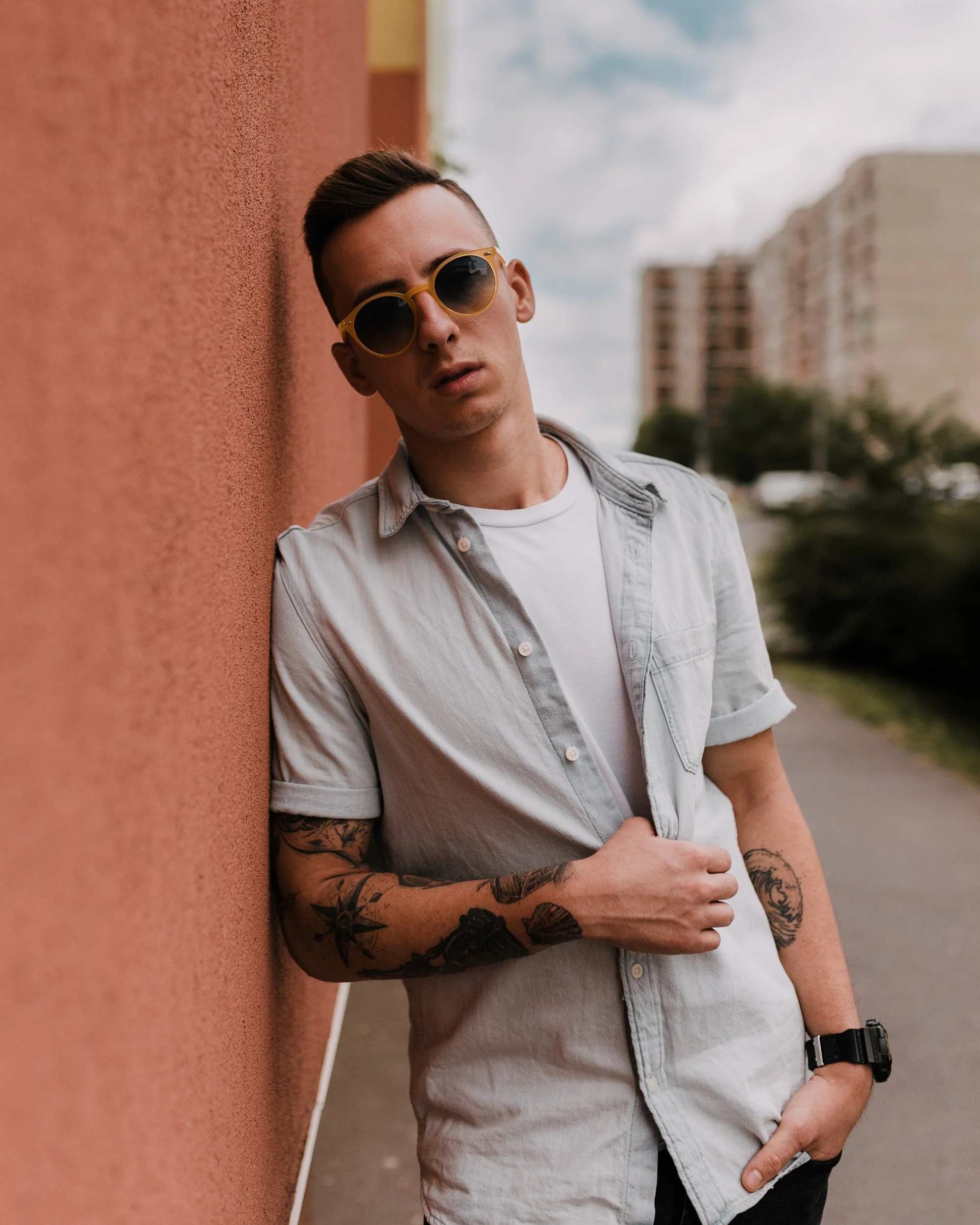 A young man with short dark hair, wearing sunglasses with yellow frames, a light-colored unbuttoned shirt over a white t-shirt, and a black watch, leaning against a pink textured wall outdoors.