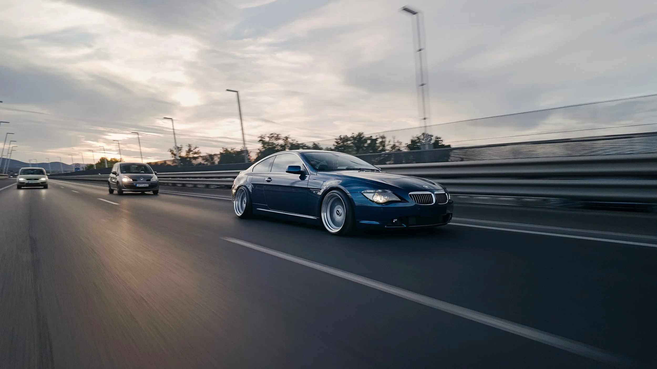 A black BMW coupe driving on a highway with other vehicles around, under a cloudy sky during sunset, with guardrails and trees beside the road.