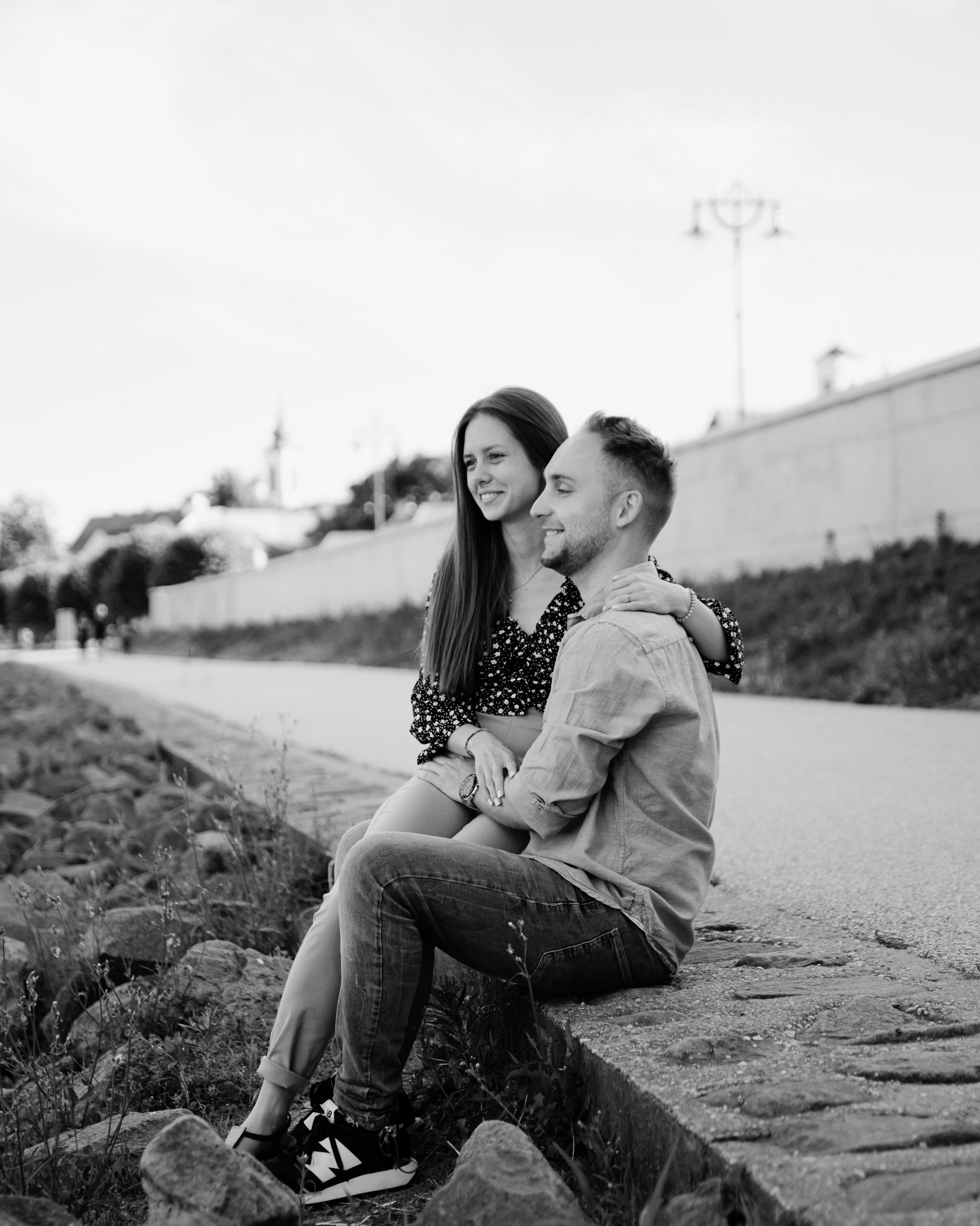 Black and white photo of a smiling couple sitting outdoors on a rock ledge, with trees and buildings in the background.
