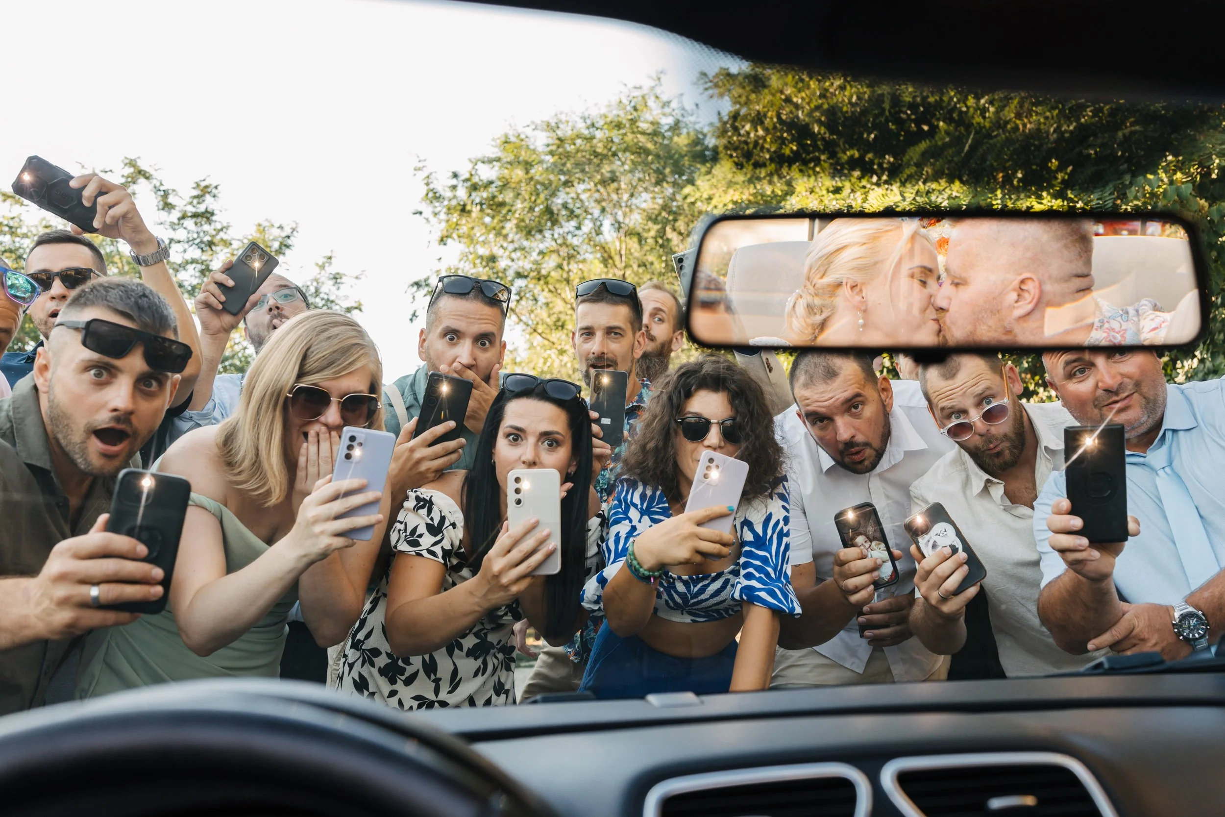 A group of people taking a selfie together outside, viewed from inside a car's windshield, with two people kissing in the rearview mirror.