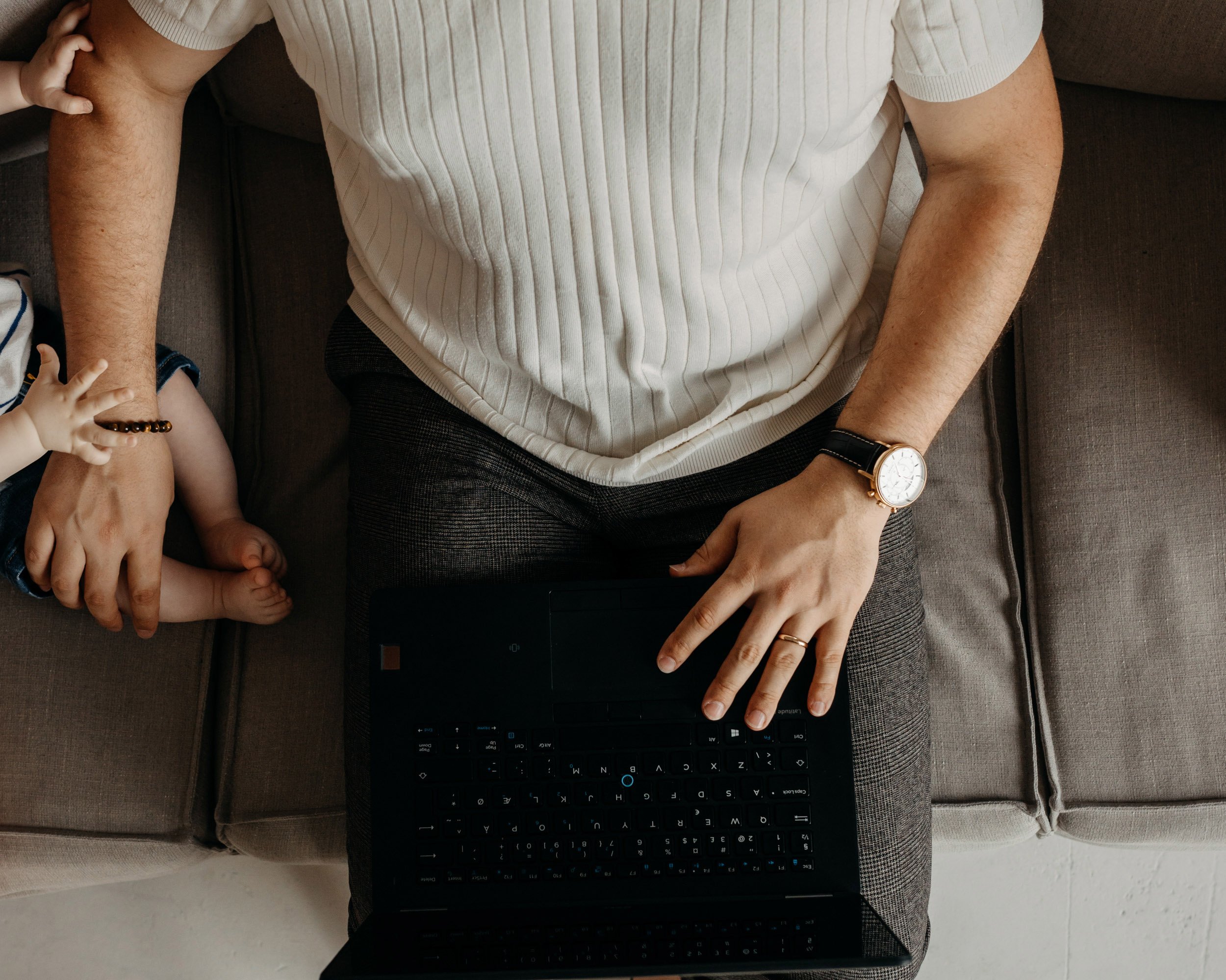 A person wearing a cream-colored, ribbed shirt sitting on a gray couch with their right arm resting on their lap and wearing a wristwatch. Part of a child's hand and arm are visible on the left side, touching the person's arm. The person is using a b