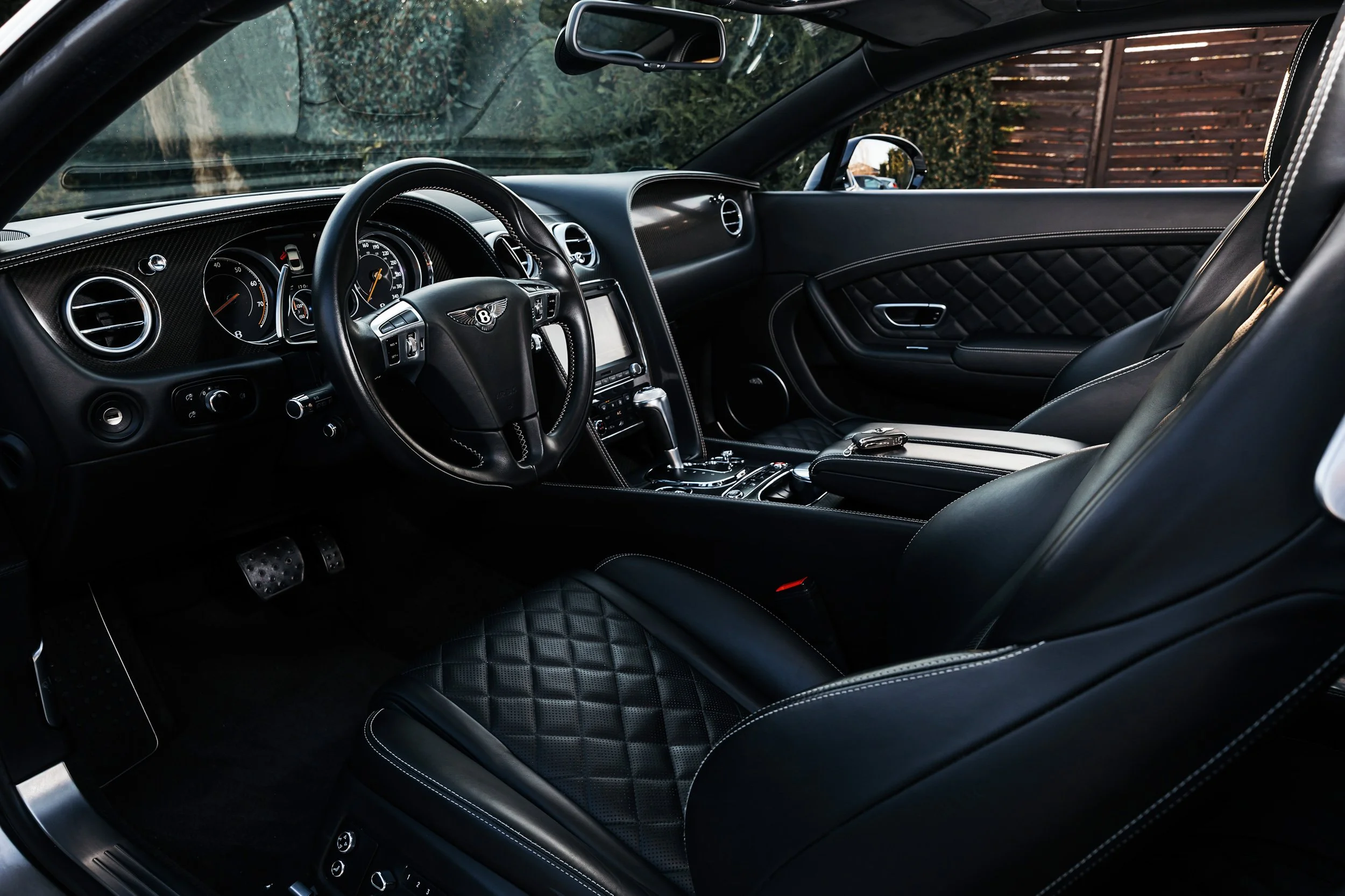 Interior of a luxury car showing the dashboard, steering wheel with Bentley logo, and black leather seats with diamond stitching.