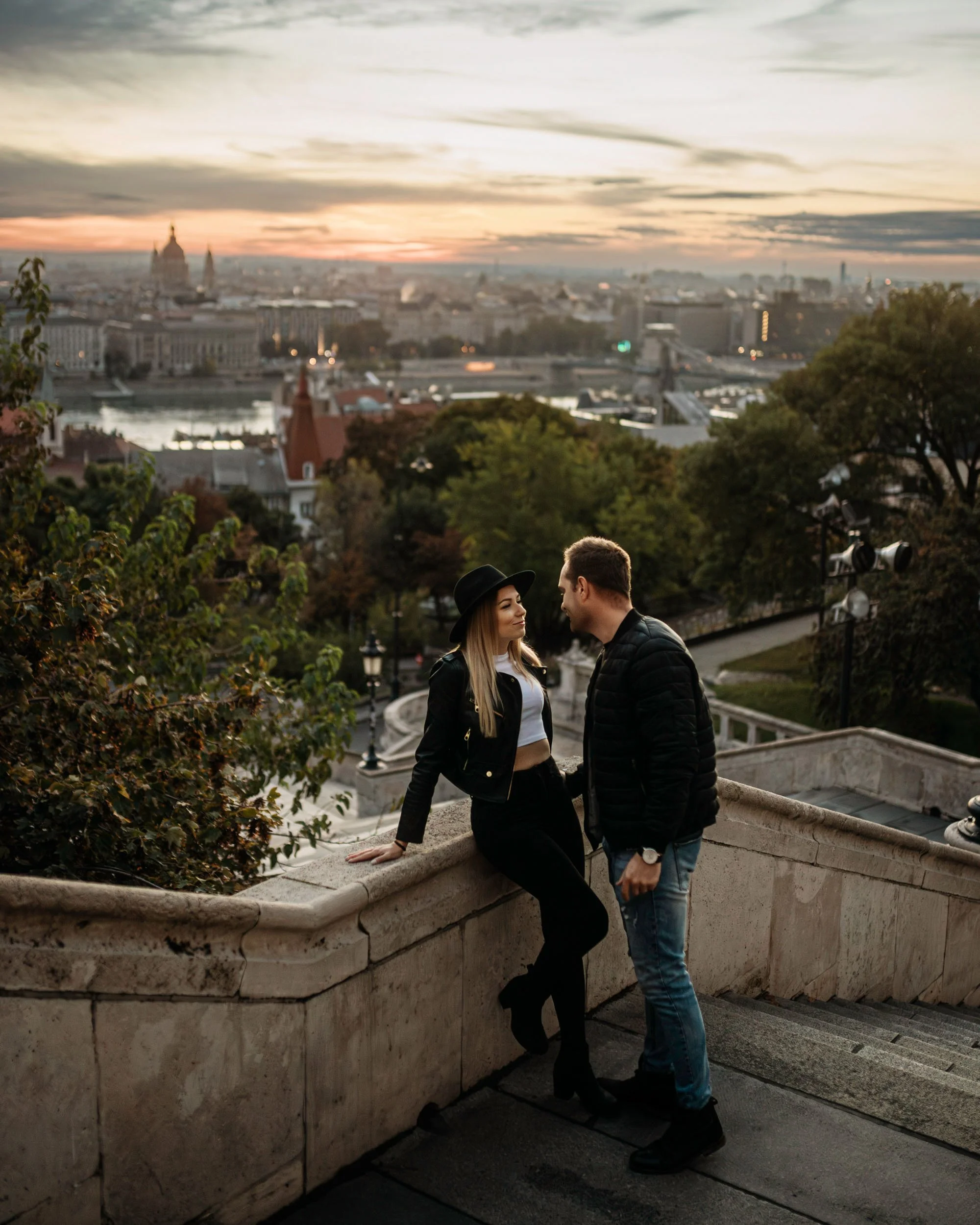 A young woman in a black hat and leather jacket sitting and smiling at a young man in a black jacket on a stone staircase overlooking a cityscape at sunset. The background shows city buildings, a river, and a cloudy sky.
