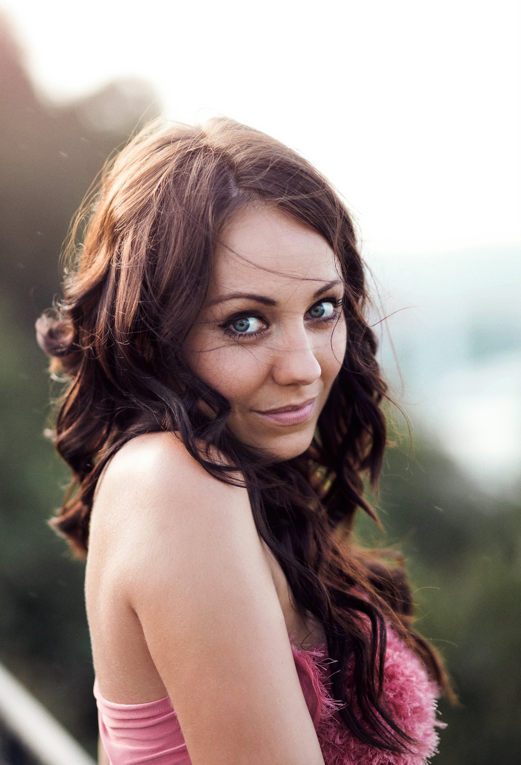 Close-up portrait of a young woman with blue eyes and long, wavy auburn hair, wearing a pink dress with a fluffy pink accessory, outdoors with a blurred natural background.
