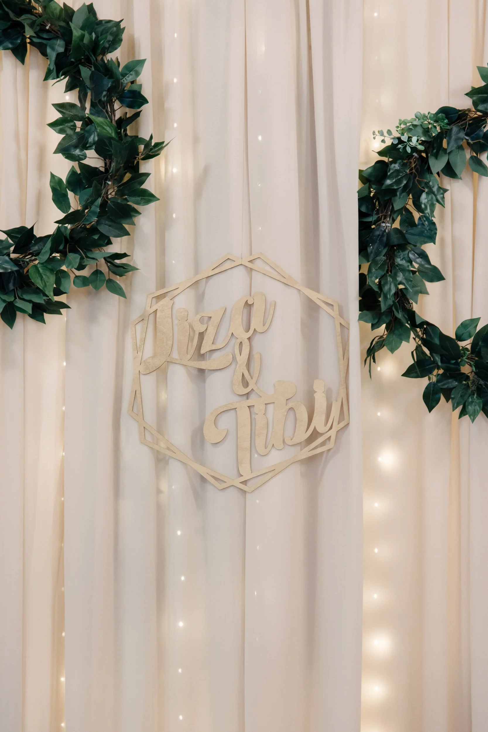 Decorative backdrop with white curtains, green leafy garlands, small string lights, and a wooden sign with the words "Wiz & Sophia".