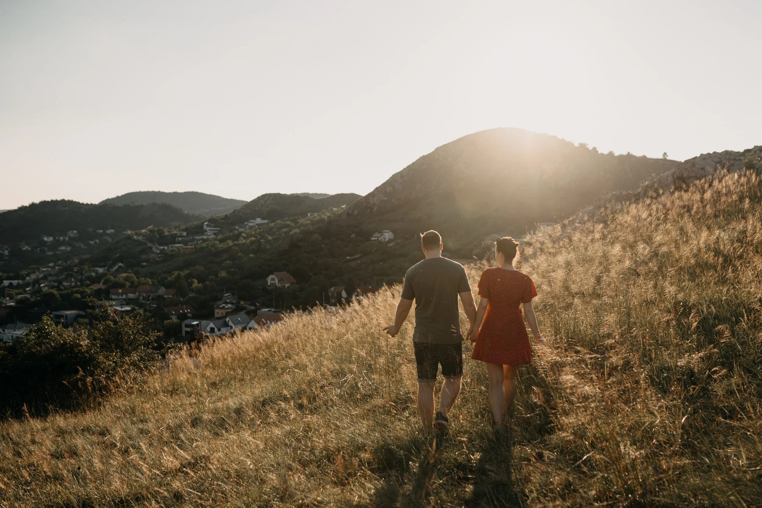 A man and a woman walk hand in hand along a grassy hillside at sunset, with a view of a hilly landscape and houses in the distance.