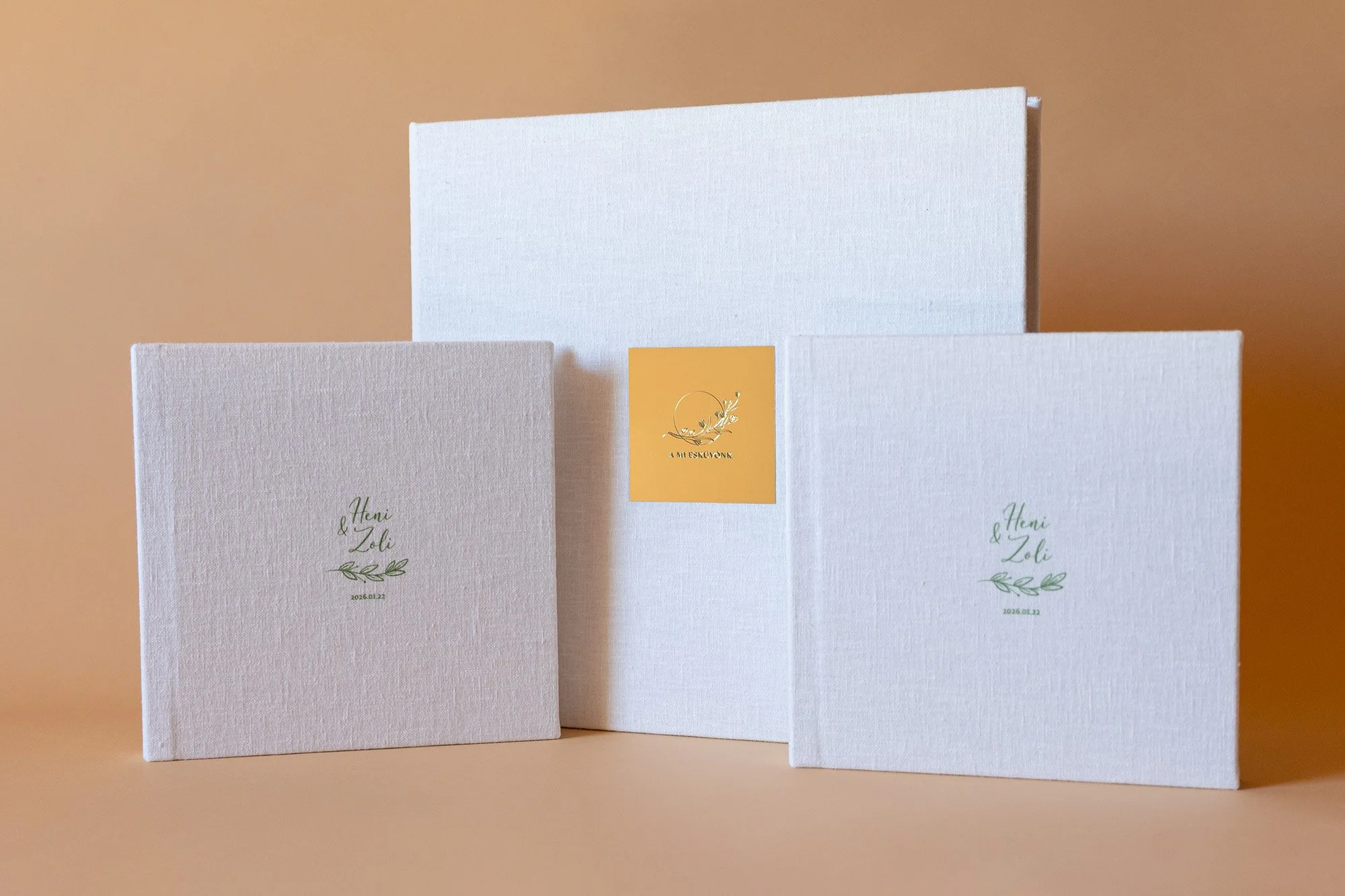 Three wedding invitation cards, two with the names 'Heni & Zoli' and a date '2026.01.22', featuring elegant script fonts and green leaf illustrations, arranged against a warm beige background.
